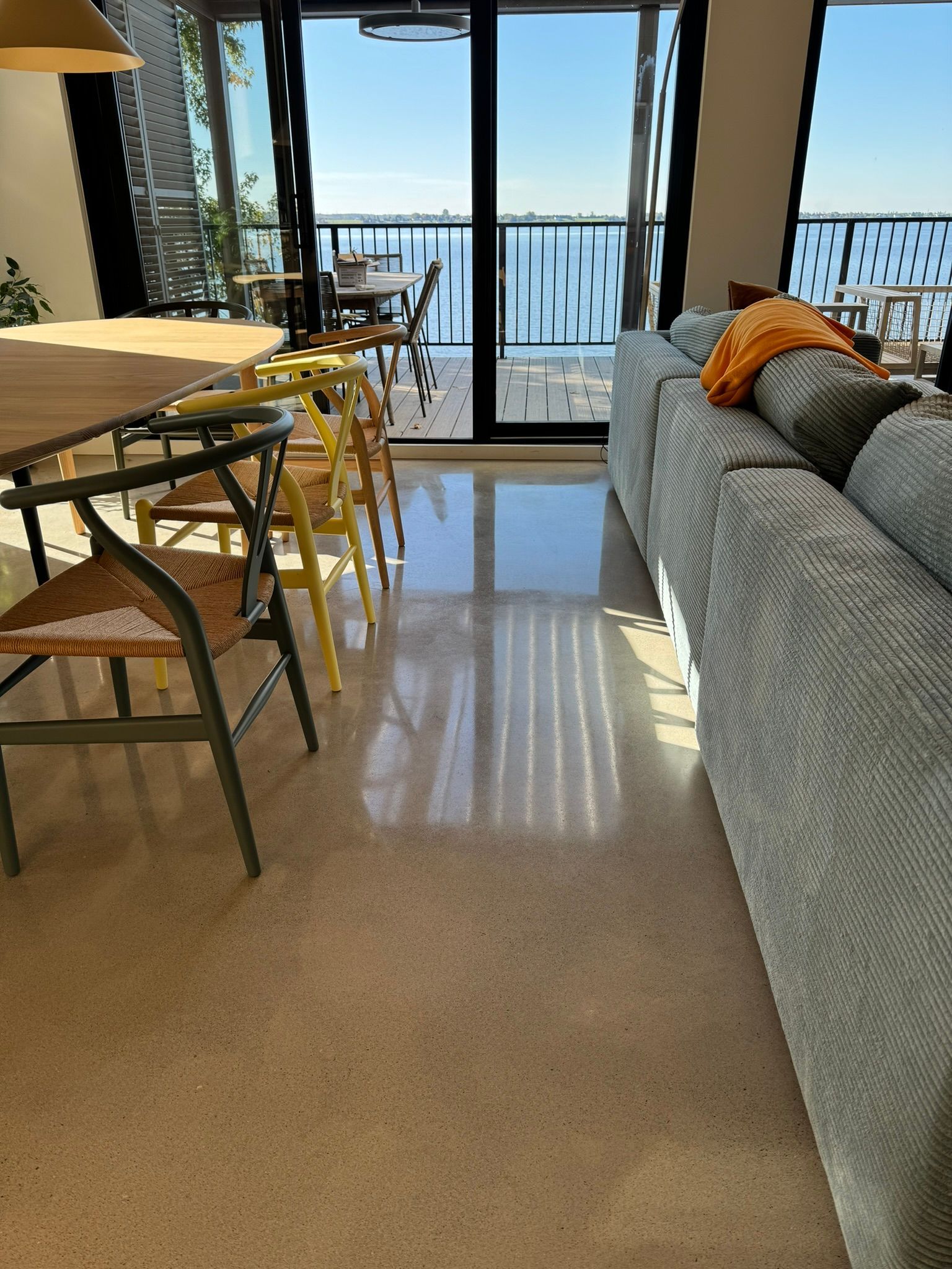 Une pièce baignée de lumière avec un sol en béton poli, un canapé gris, une table à manger et une vue sur l'océan à travers une porte vitrée.
