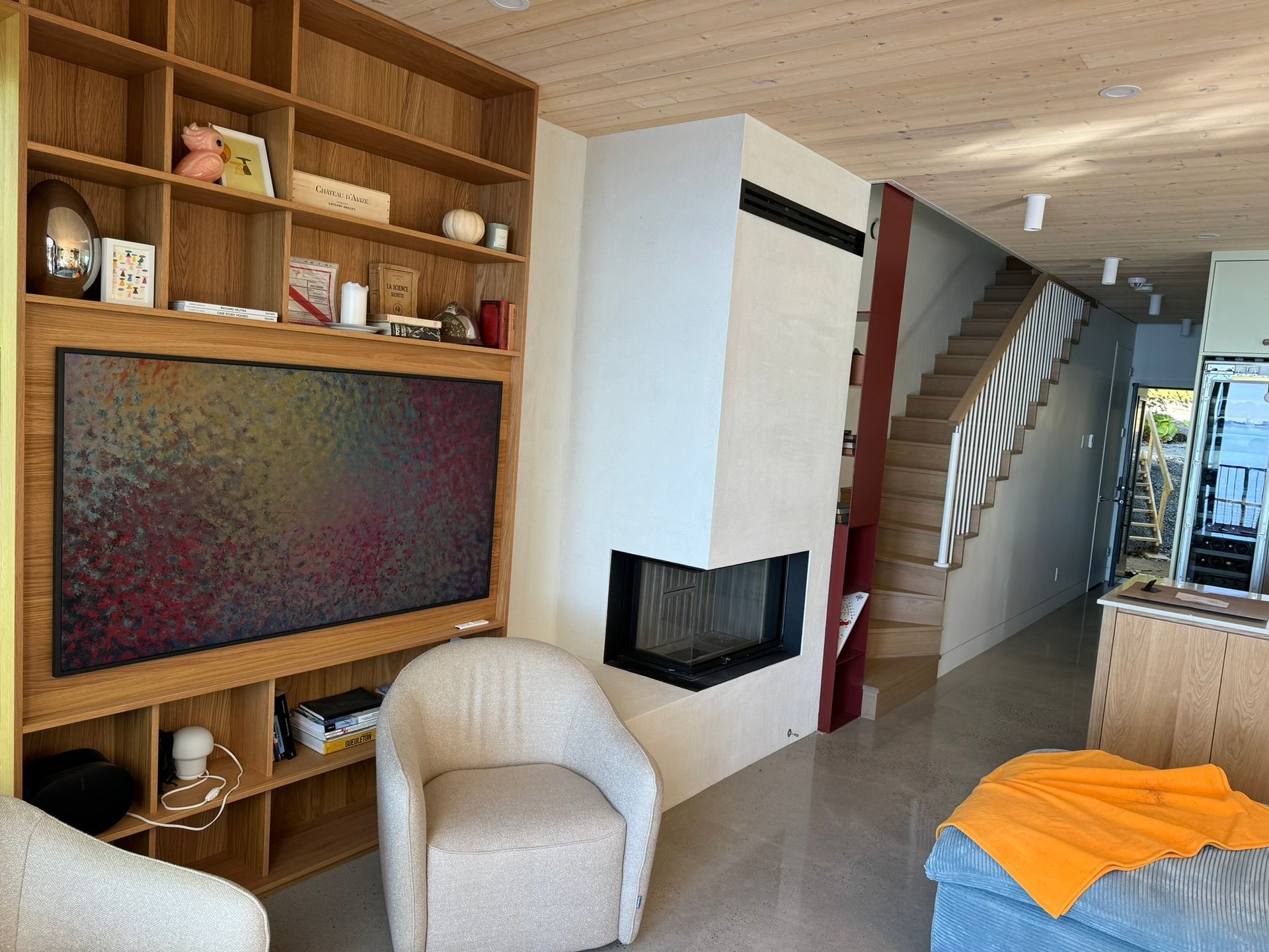 Salon avec étagères en bois intégrées, grande télévision, cheminée, escalier et fauteuil blanc.