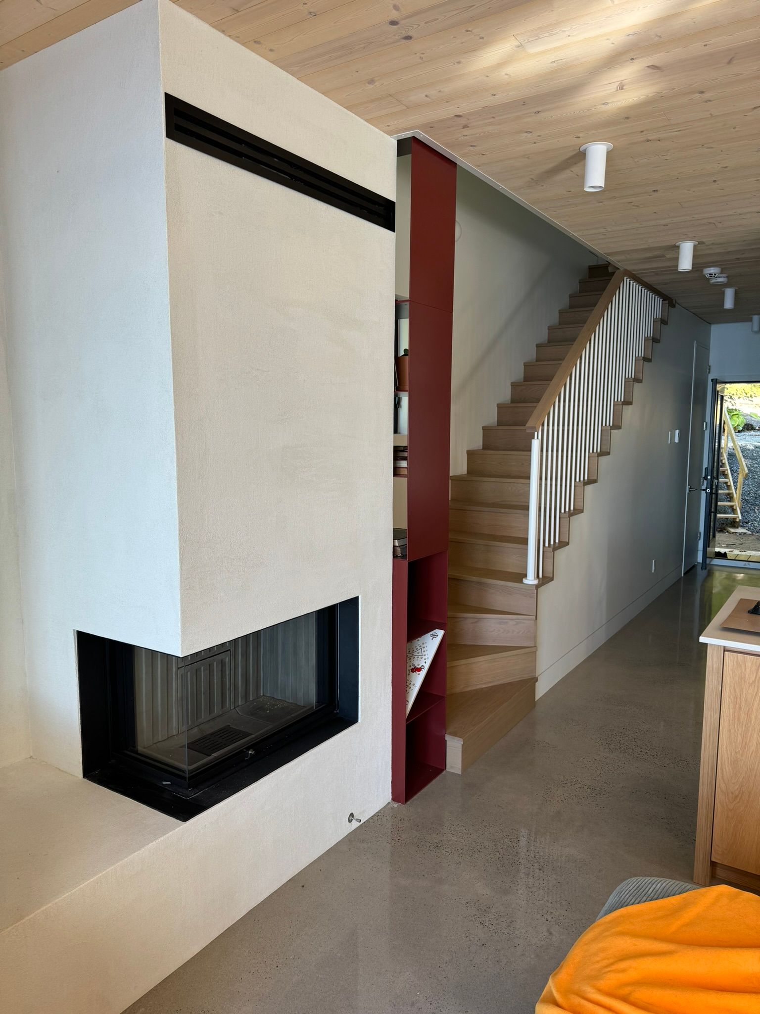 Cheminée, escalier et couloir intérieur. Murs blancs et rouges, escalier en bois, sol en béton, éclairage au plafond.