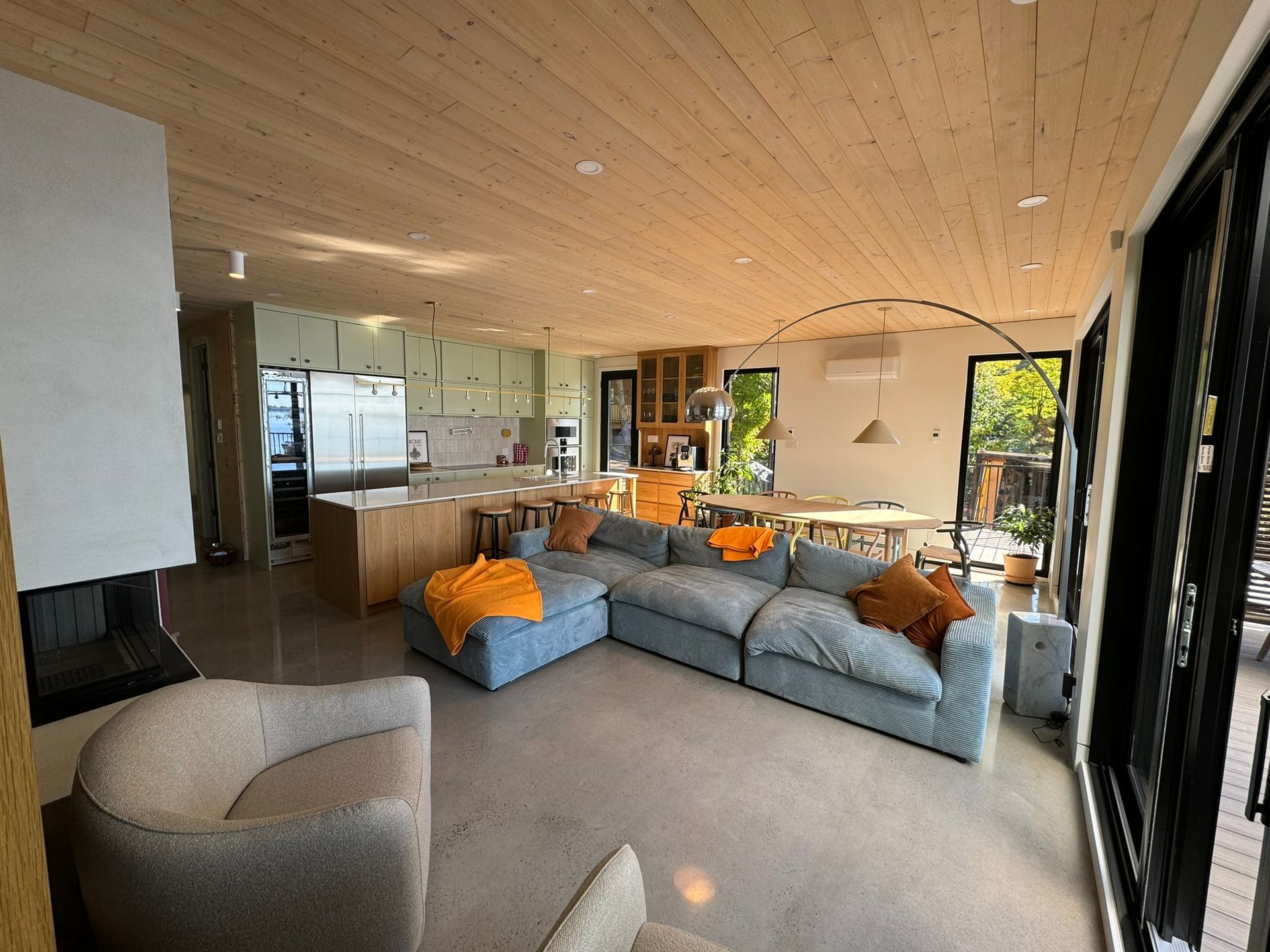 Espace de vie moderne et ouvert avec plafond en bois clair, canapé gris, grandes fenêtres et fauteuil beige incurvé.