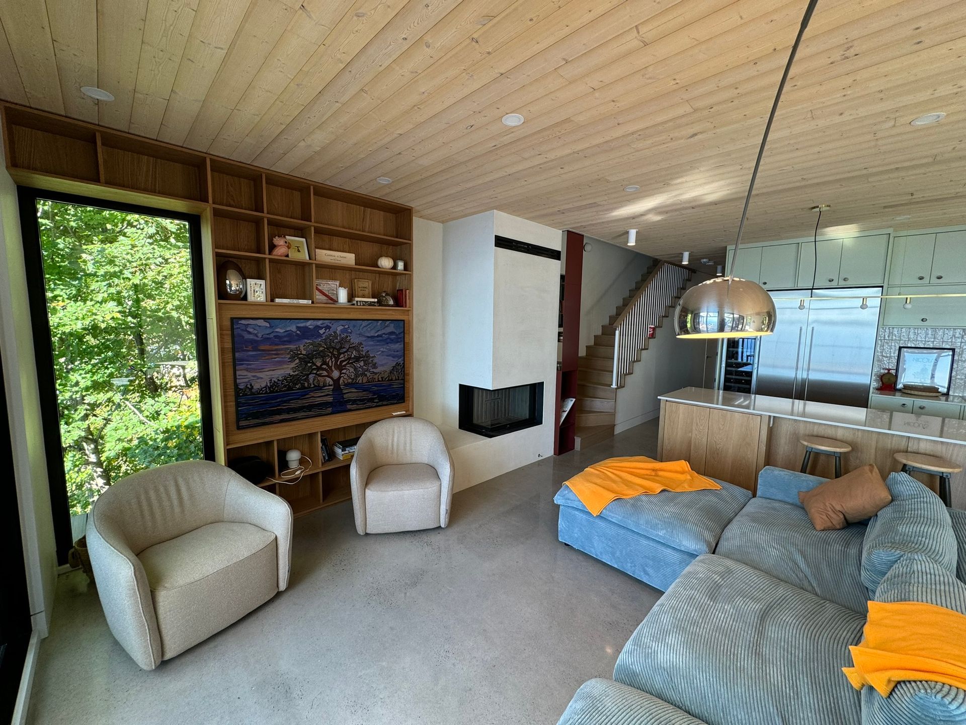 Salon chaleureux avec cheminée, étagères intégrées, canapé bleu et fauteuils. De grandes fenêtres offrent une vue sur les arbres.