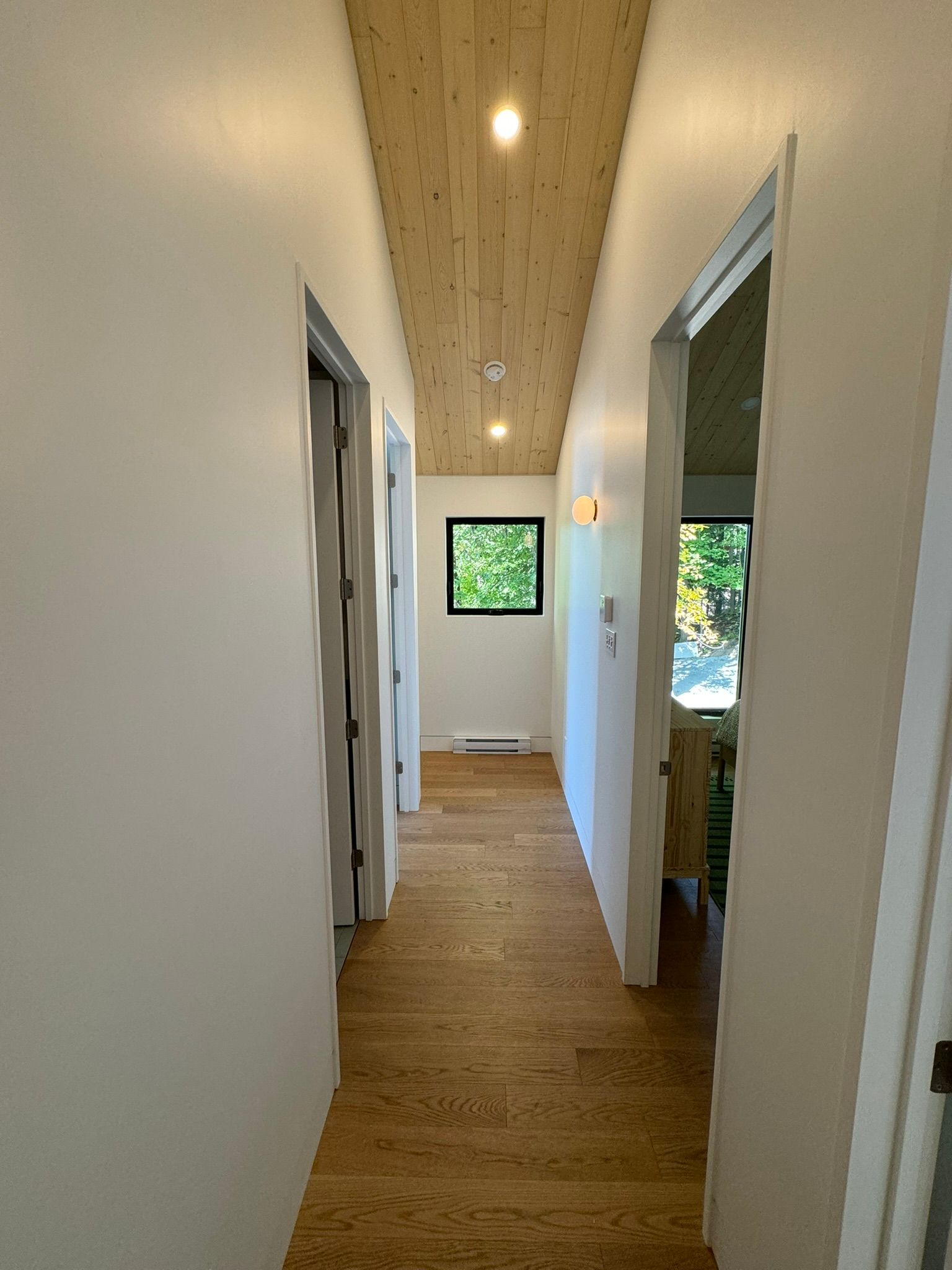 Couloir étroit avec sol et plafond en bois, murs blancs, portes et une petite fenêtre au fond.