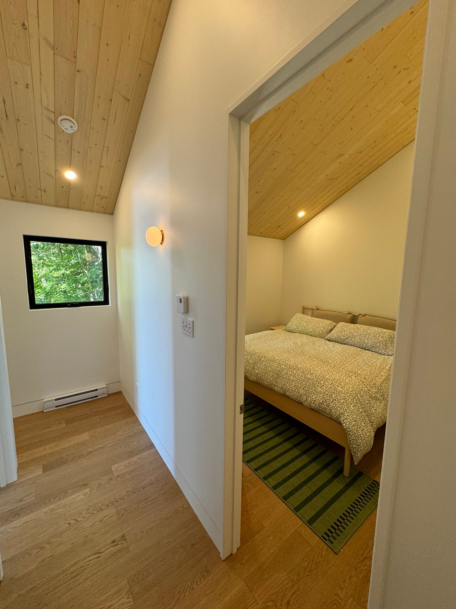 Vue intérieure d'une chambre avec parquet et plafond en bois, porte donnant sur une pièce avec un lit et un couloir.