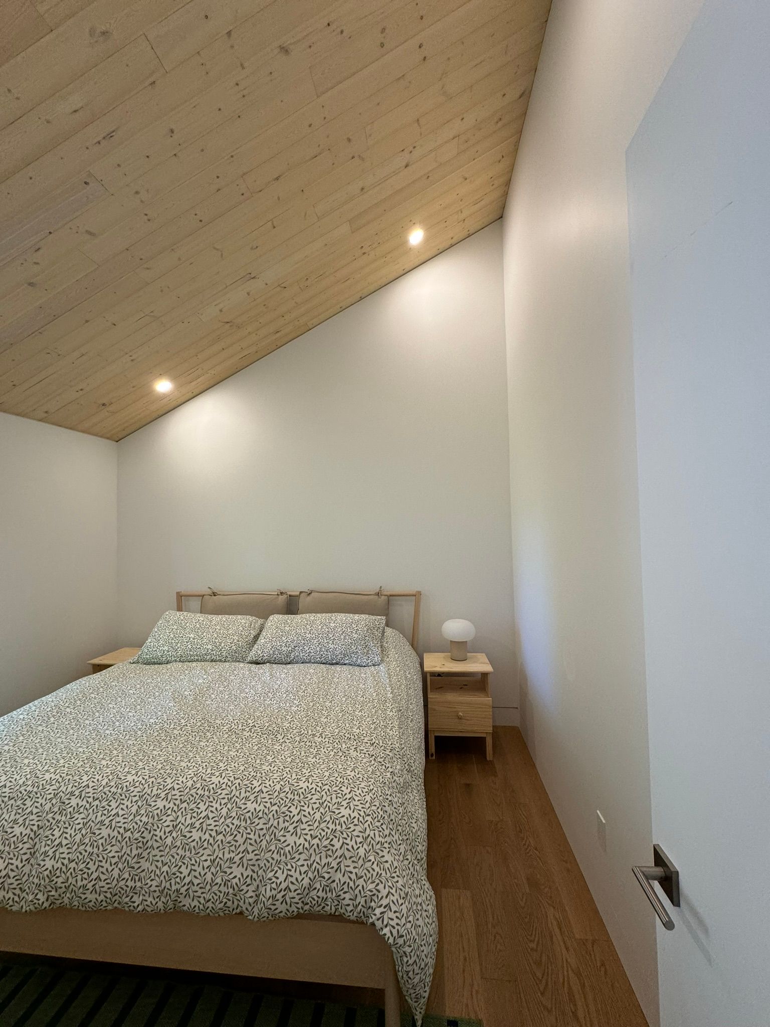 Chambre avec plafond en bois, murs blancs, lit recouvert d'une couverture à motifs et parquet.