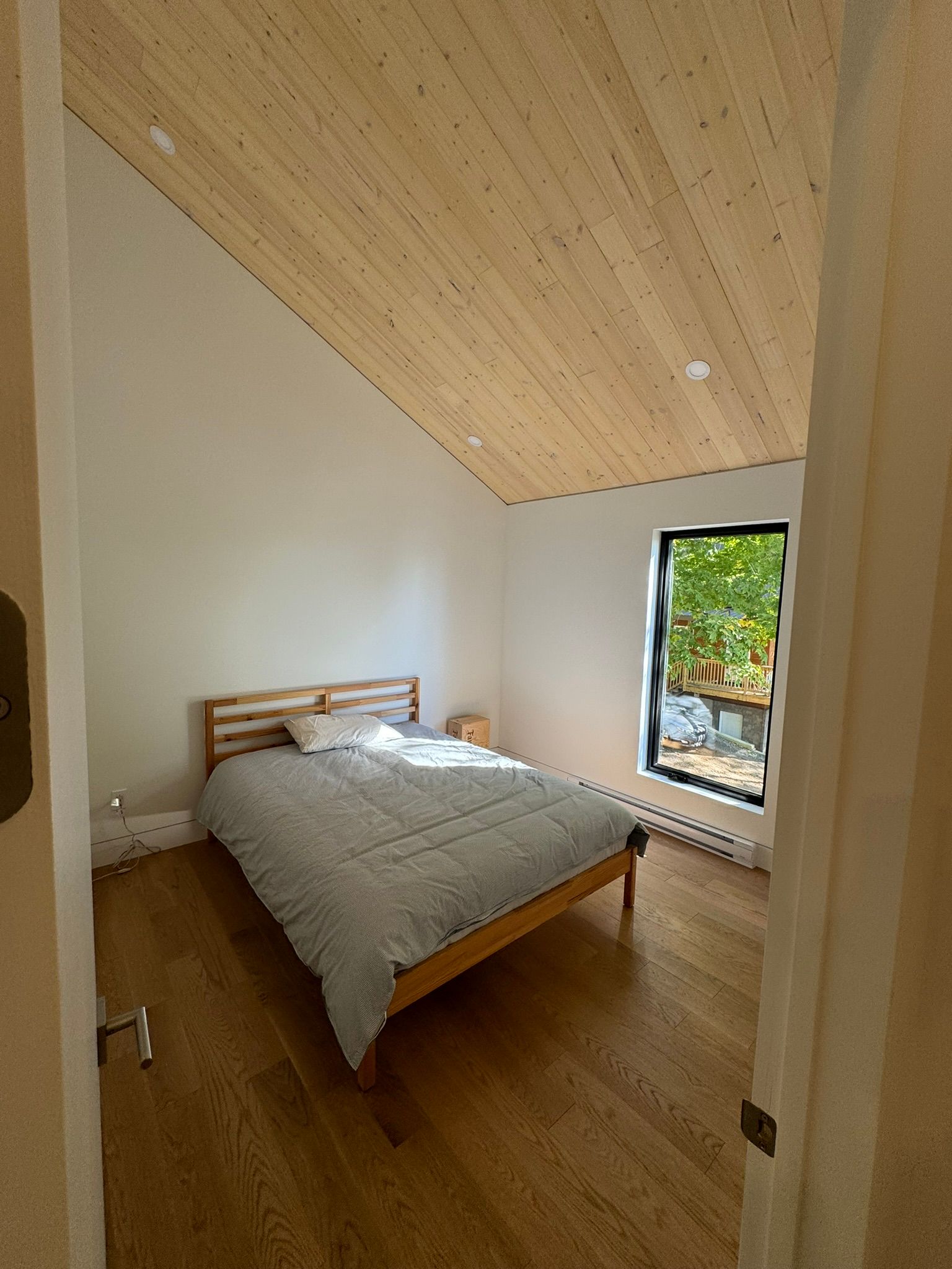 Chambre avec parquet, plafond en pente en bois, lit et fenêtre donnant sur l'extérieur.