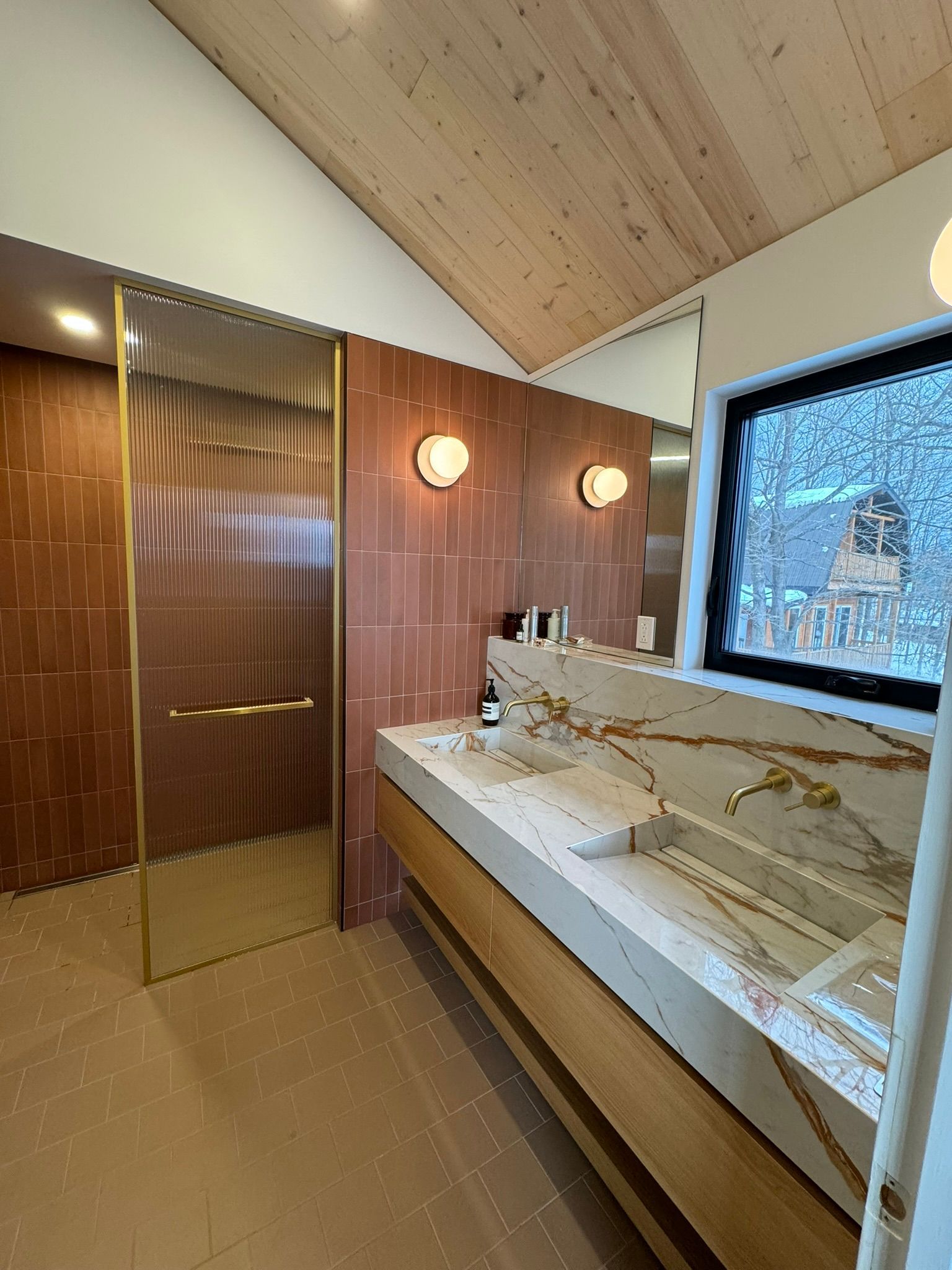 Salle de bains avec carrelage rouge, touches dorées, plafond en bois et long comptoir à motifs.