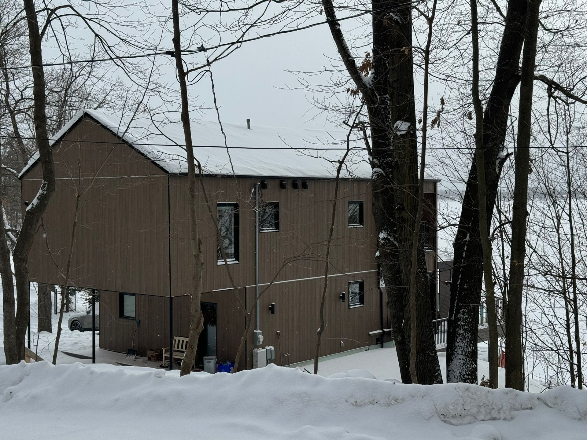 Maison moderne brune dans une zone boisée et enneigée. Fenêtres, toit sombre, neige au sol.