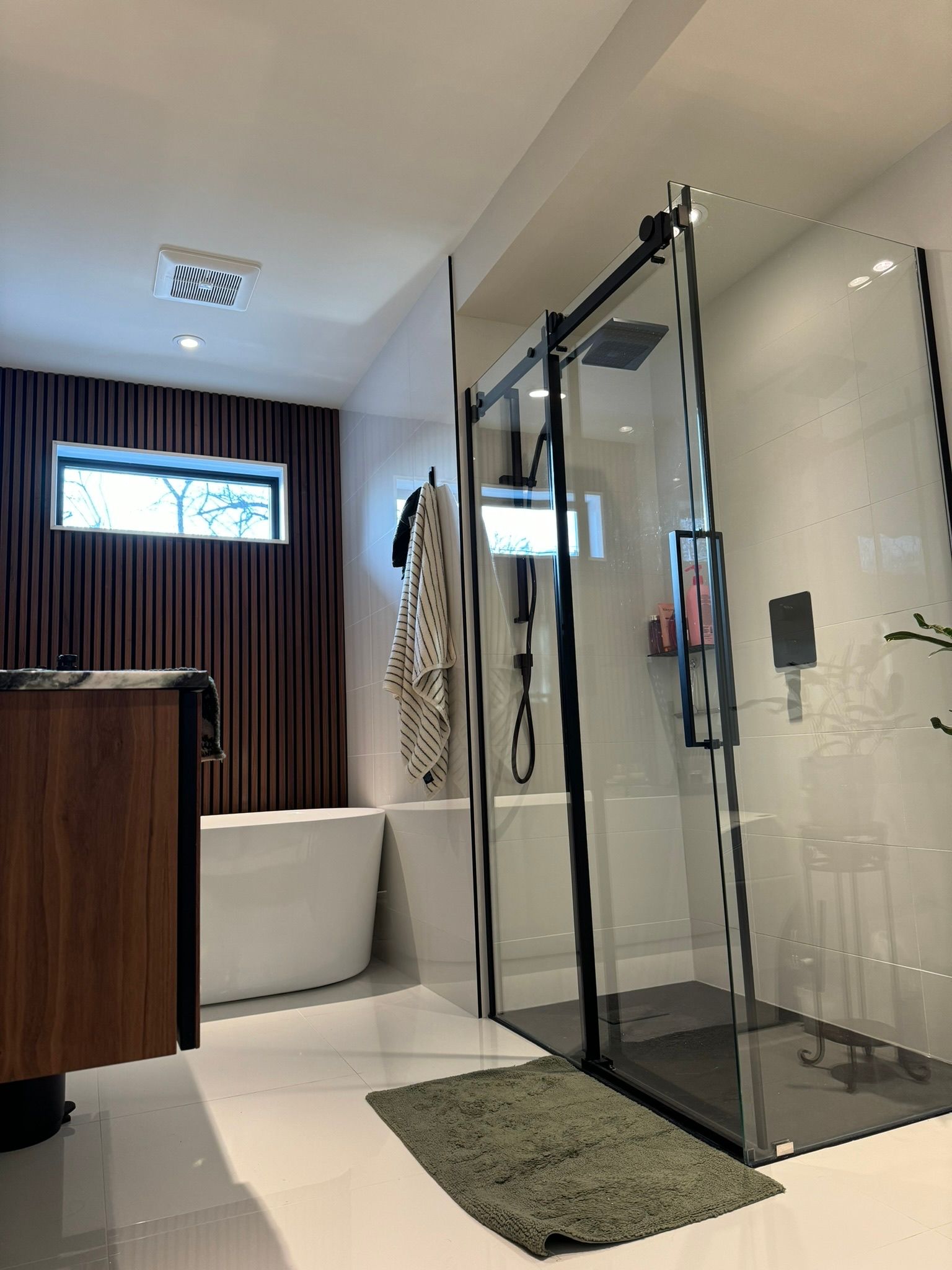Salle de bains moderne avec douche en verre, baignoire, meuble-lavabo en bois et mur d'accent texturé marron.