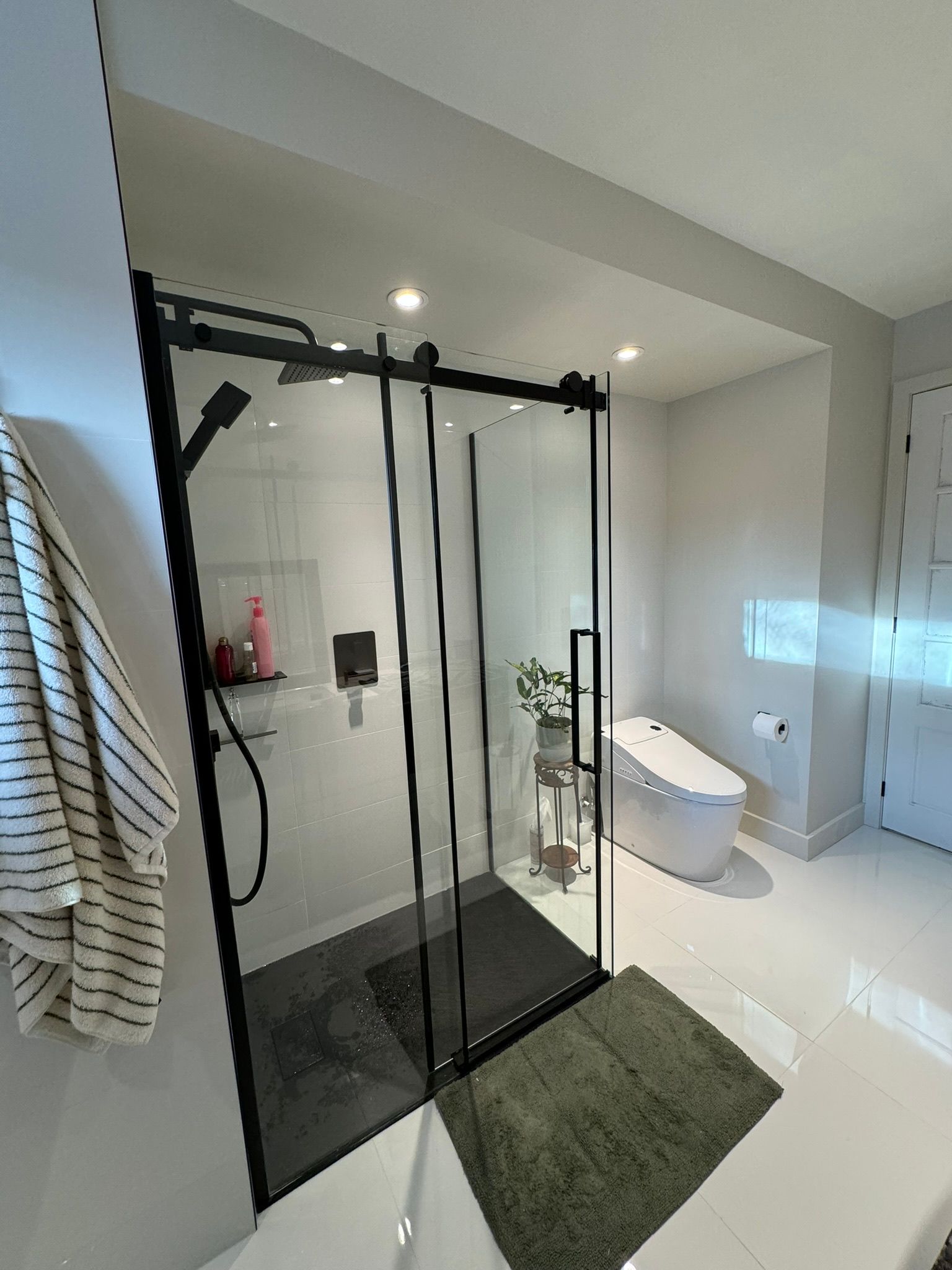 Salle de bains moderne avec douche à l'italienne encadrée de verre noir. Toilettes blanches, tapis vert et murs gris.