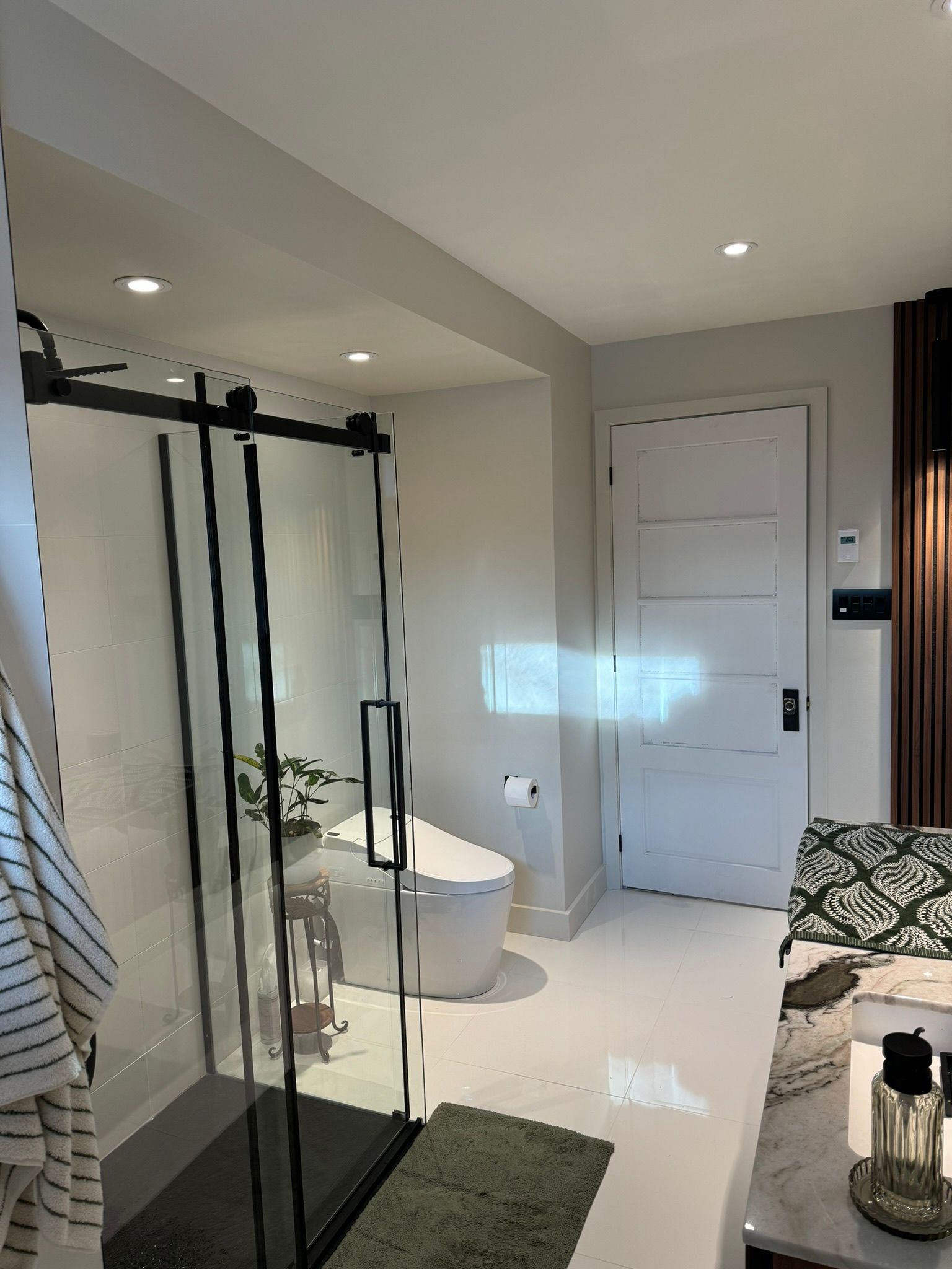 Salle de bains moderne avec douche en verre, toilettes blanches, porte et tapis décoratif.