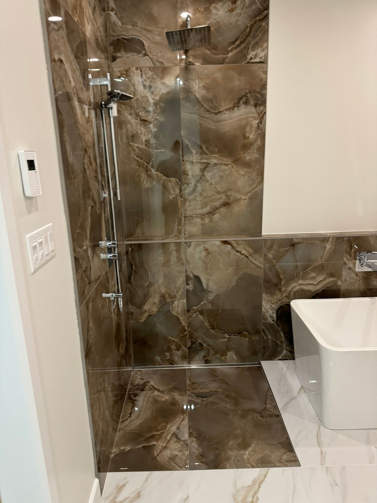 Douche avec carrelage imitation marbre brun, porte en verre et robinetterie chromée. Une baignoire blanche se trouve juste à côté.
