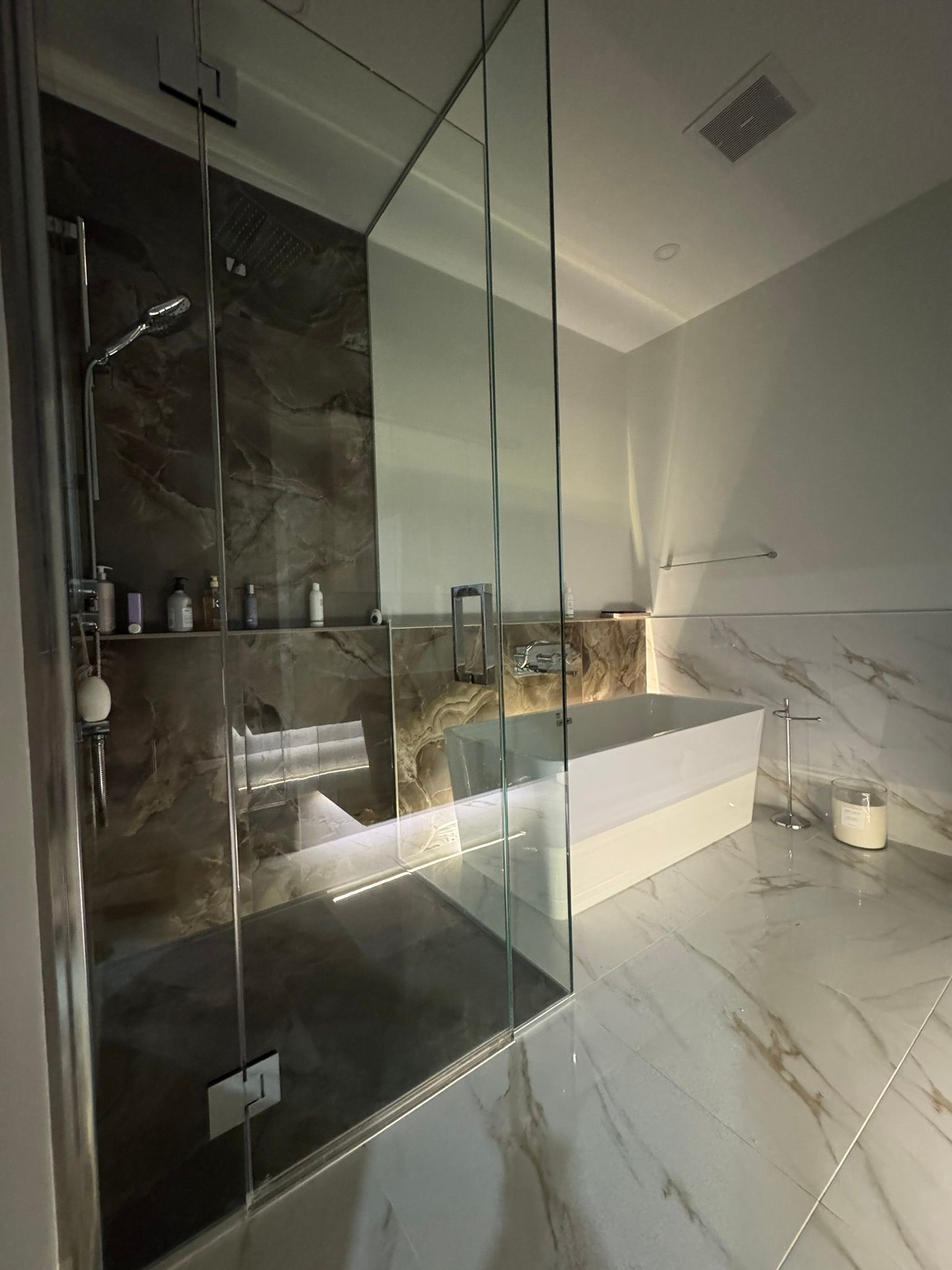 Salle de bains moderne avec douche en verre, baignoire, murs et sol en marbre et éclairage encastré.