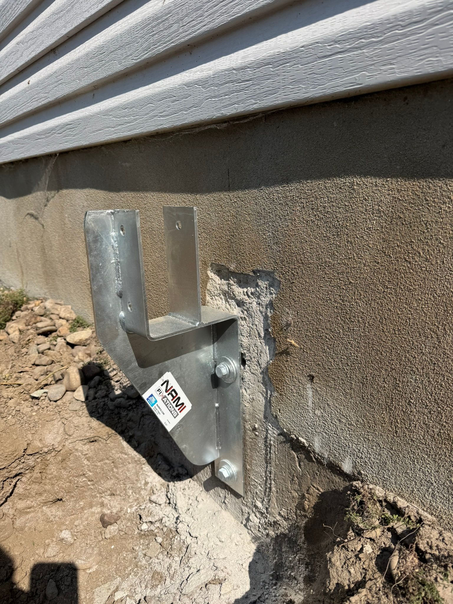 Un support métallique est boulonné à une fondation en béton. La terre entoure le support. Le revêtement extérieur de la maison se trouve au-dessus.