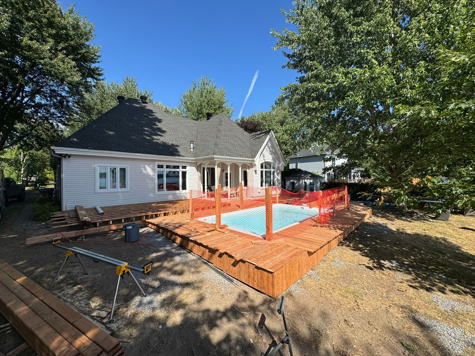 Maison avec une terrasse en bois neuve entourant une piscine rectangulaire en construction. Clôture de sécurité orange.