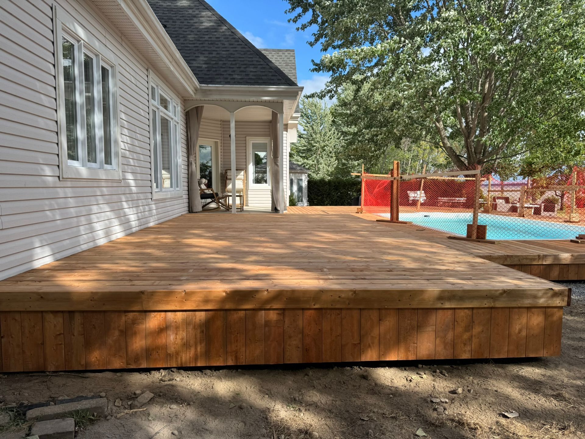 Terrasse en bois prolongeant une maison blanche. Piscine avec clôture orange en arrière-plan.