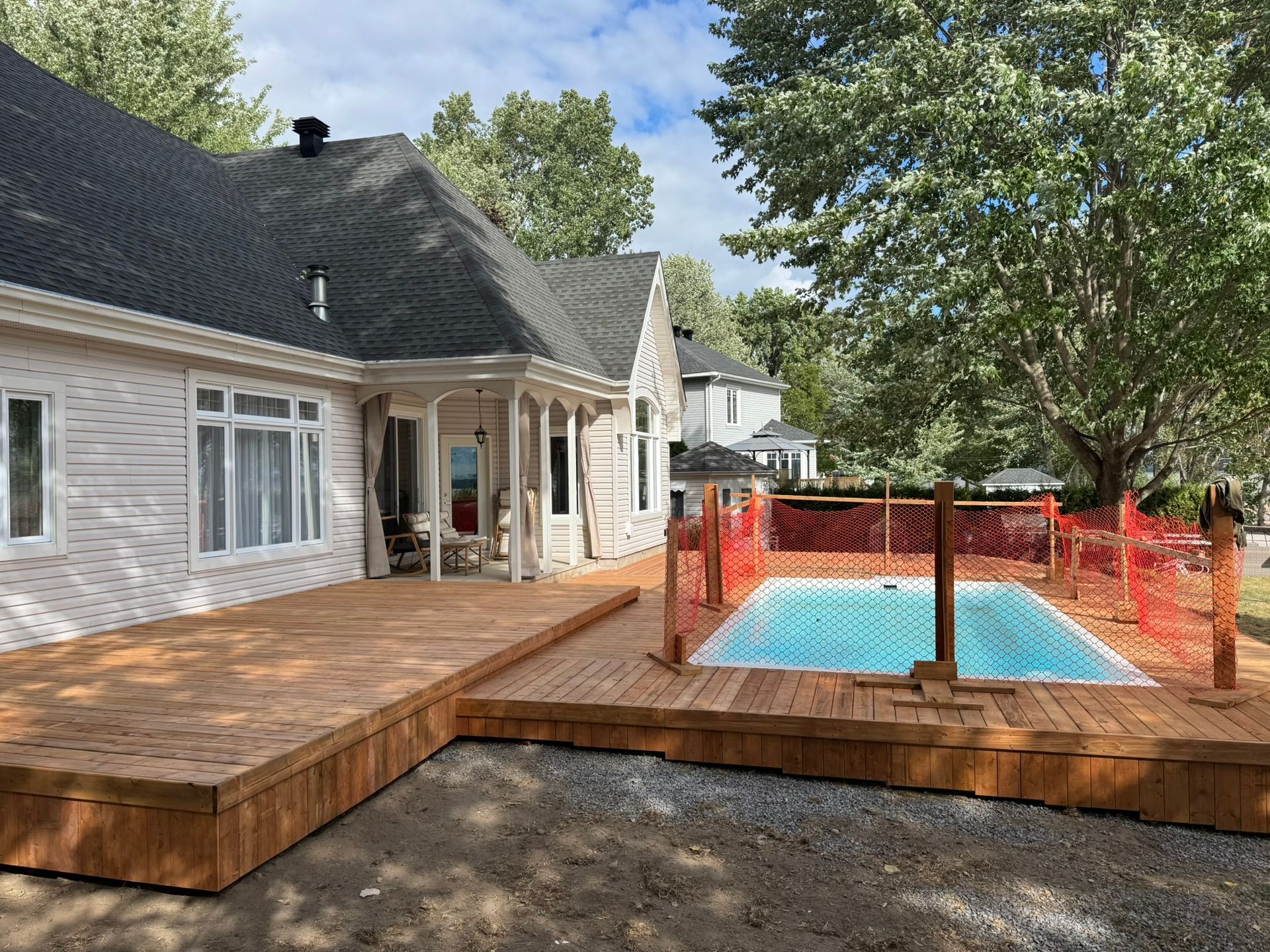 Une terrasse en bois entoure une petite piscine clôturée, à côté d'une maison aux couleurs claires.
