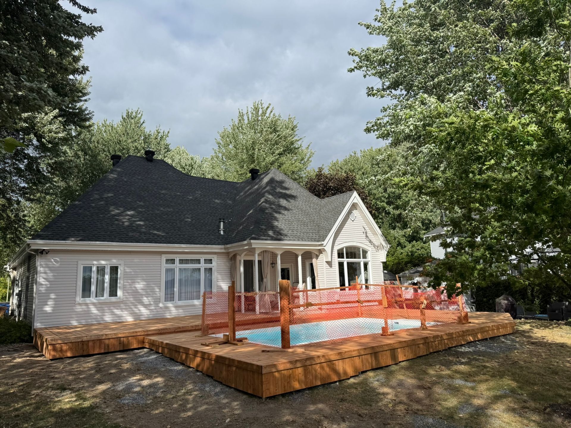 Maison avec terrasse en bois et piscine, entourée d'arbres. Une clôture de sécurité orange entoure la piscine.