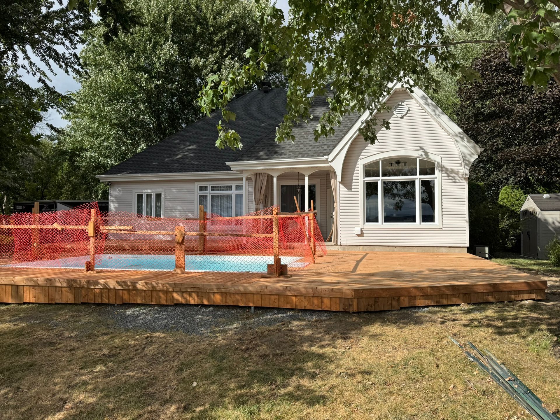 Petite maison claire avec terrasse en bois et piscine clôturée. Arbres en arrière-plan.