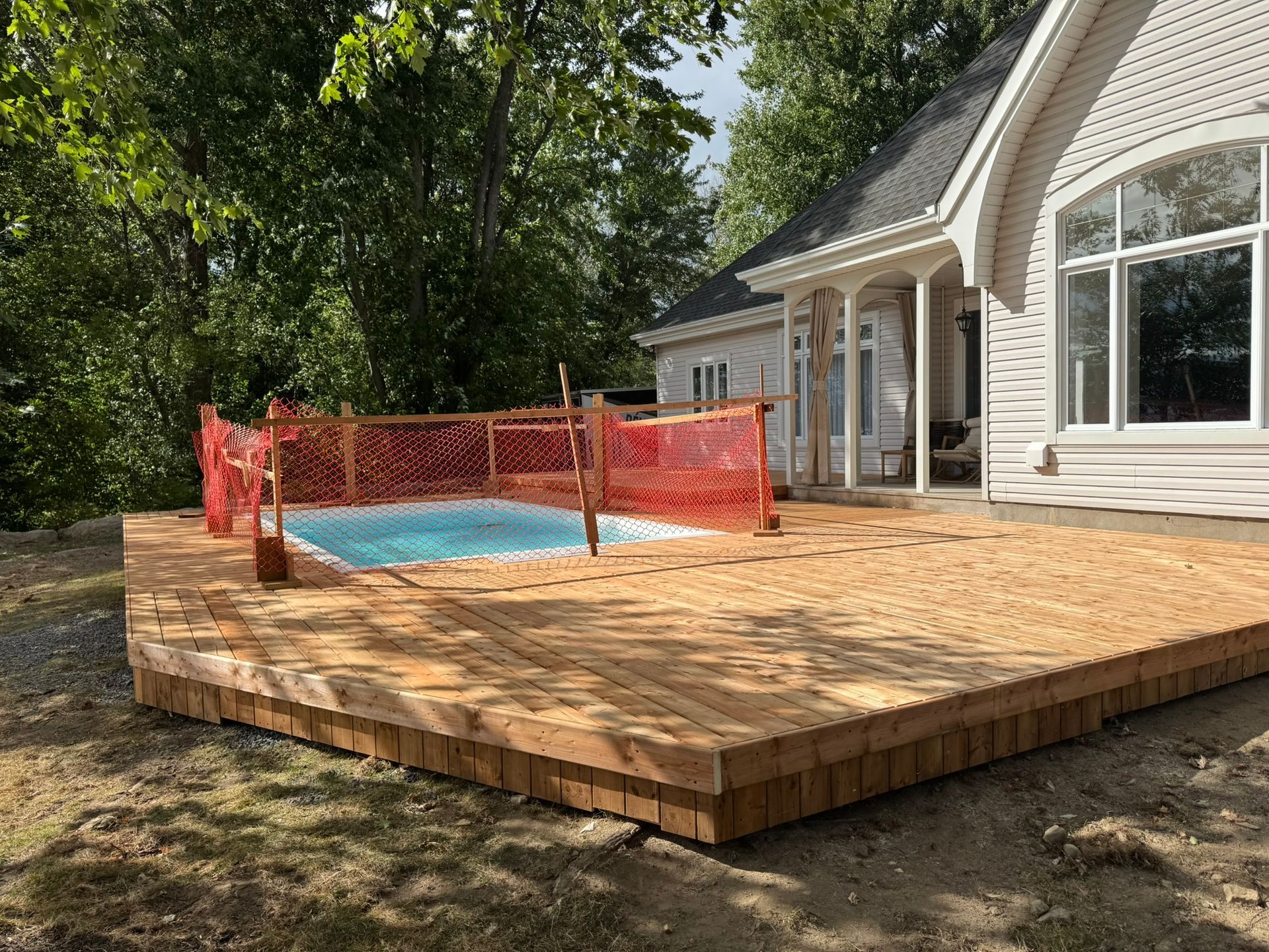 Terrasse en bois avec petite piscine, barrière de sécurité orange, près d'une maison aux grandes fenêtres.