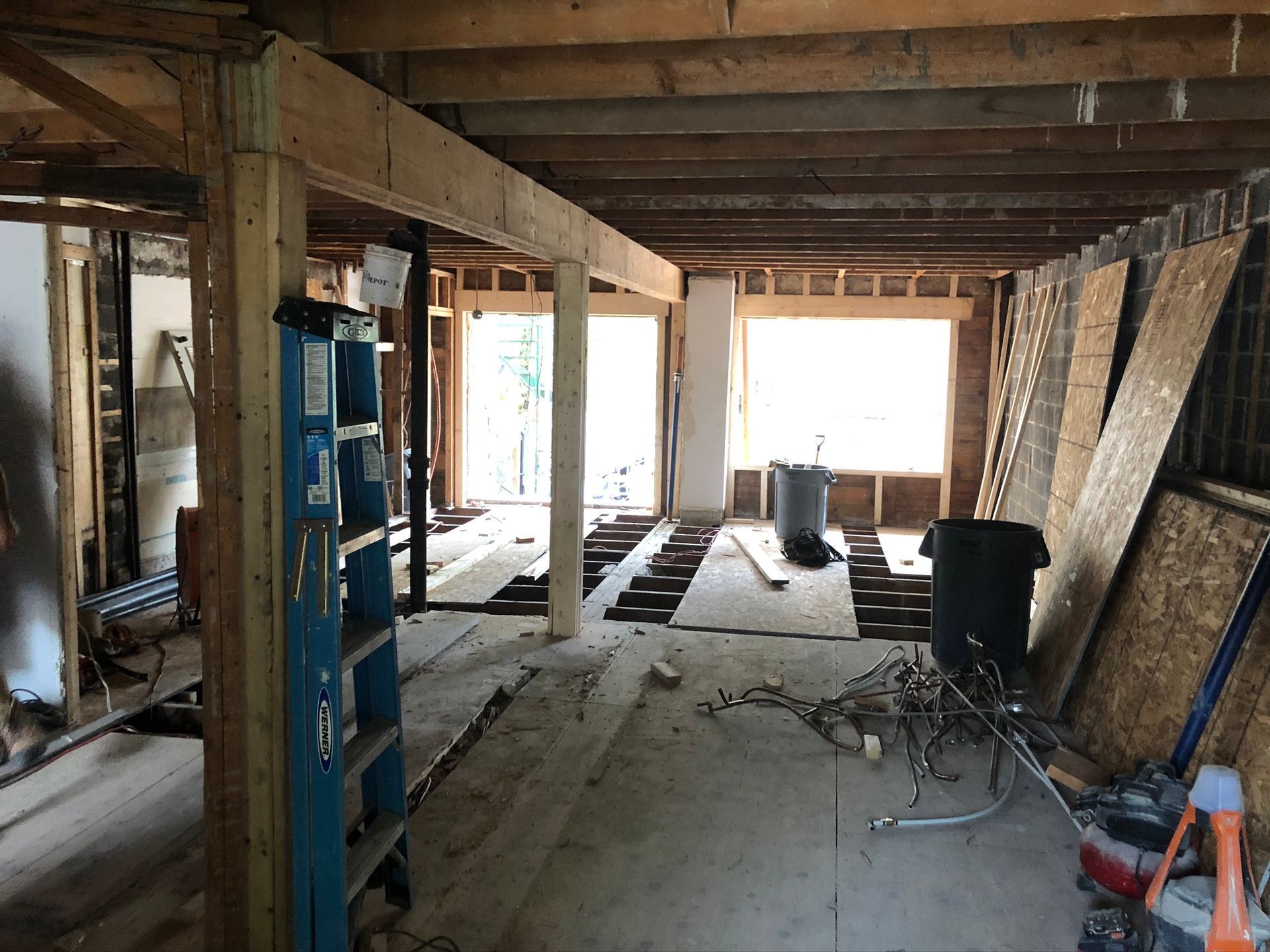 Rénovation intérieure en cours. Montants, poutres et plancher apparents ; une échelle bleue ; débris et matériaux éparpillés.