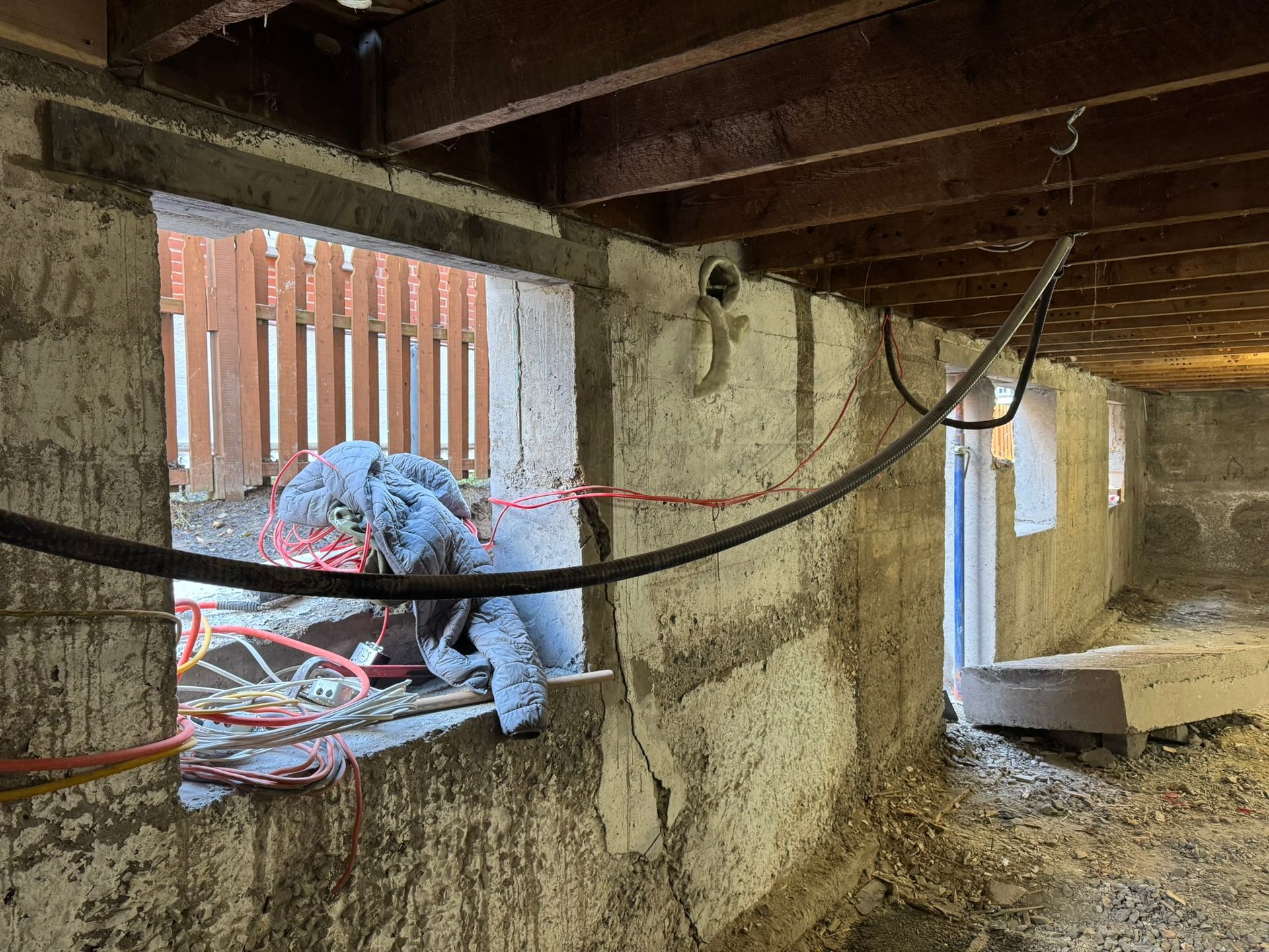 Vue intérieure d'un sous-sol en construction avec poutres et câbles apparents, et une fenêtre donnant sur une clôture en bois.