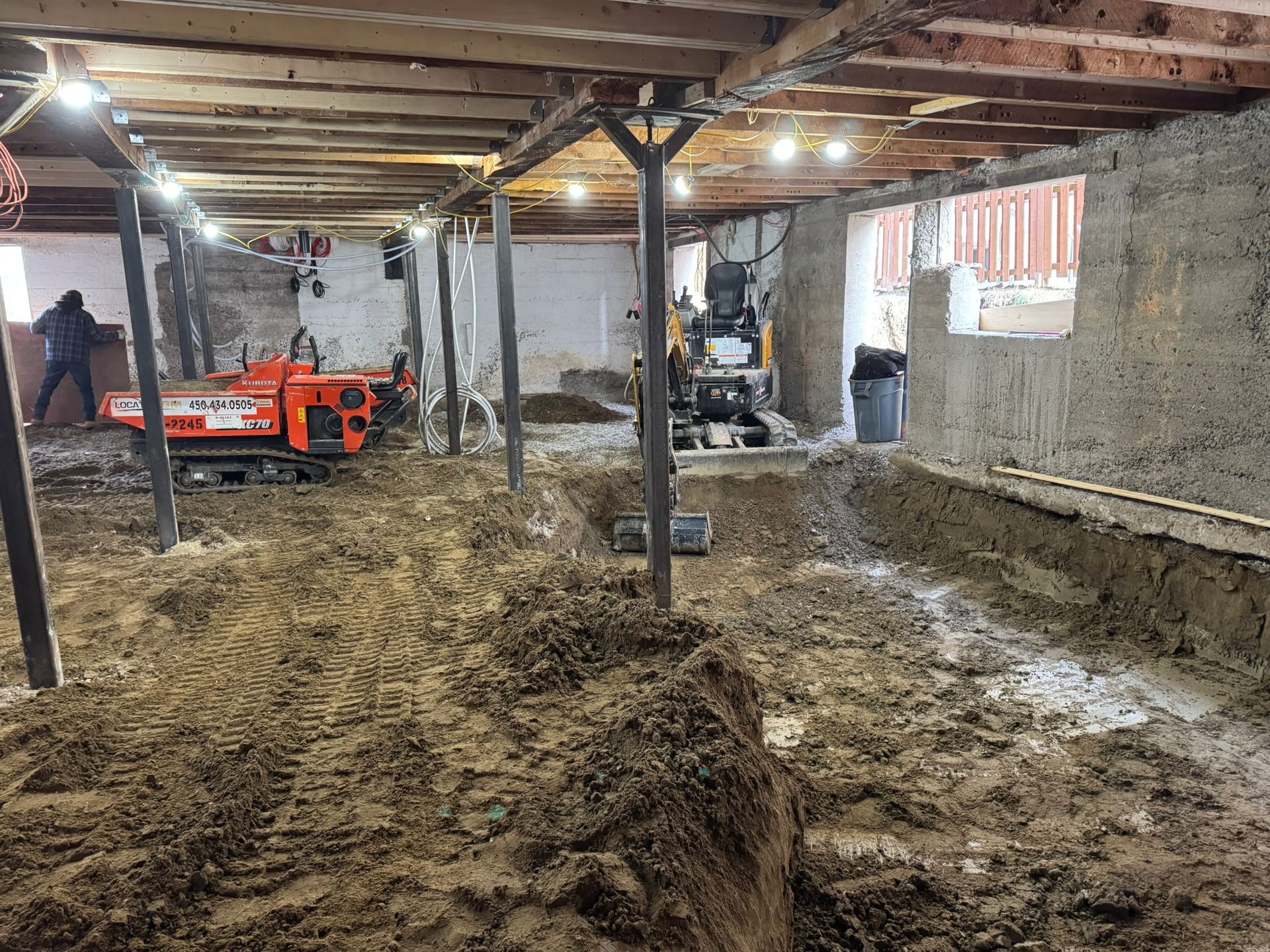 Sous-sol boueux en construction avec poutres, équipements et ouvrier.
