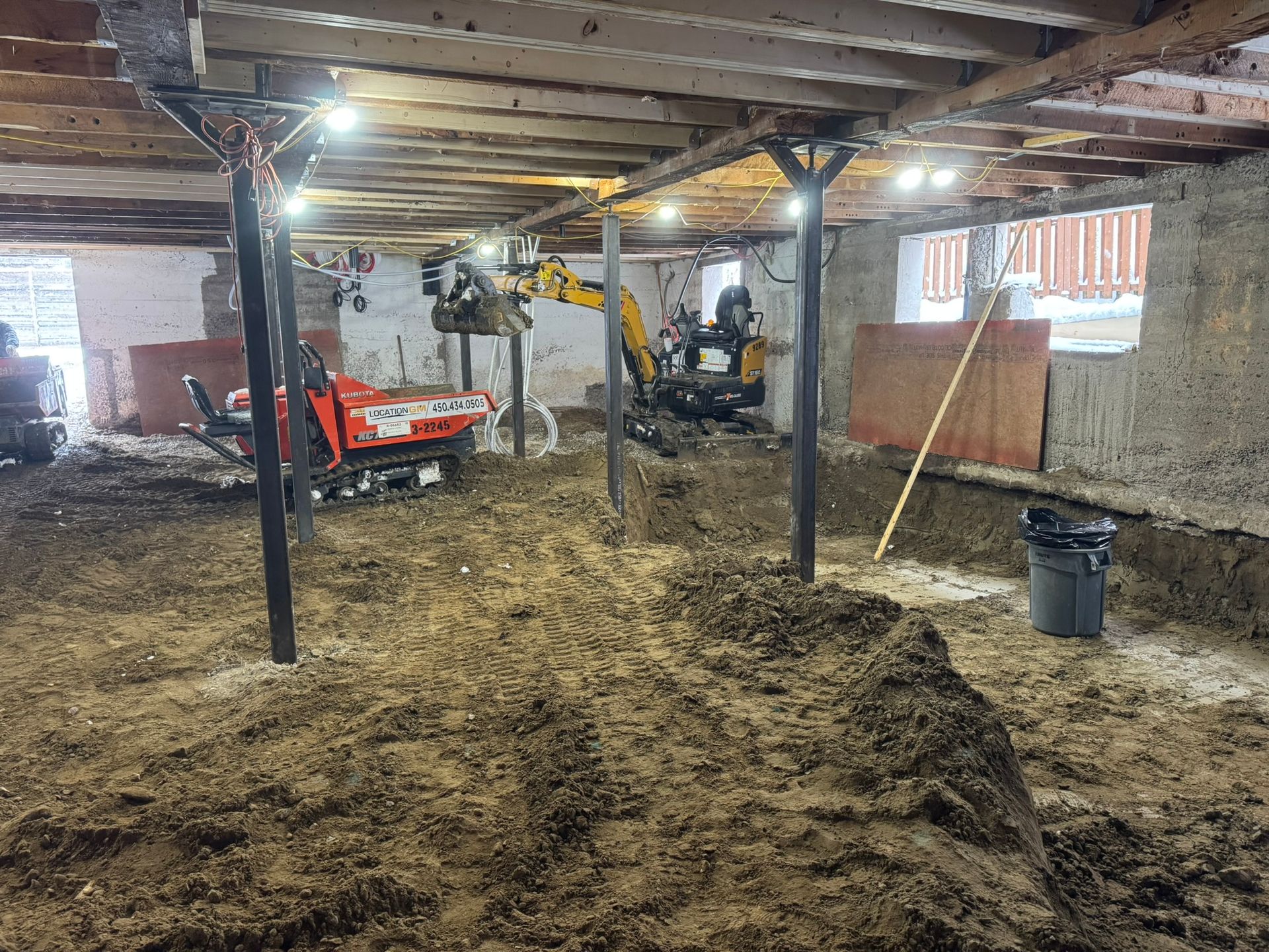 Chantier en sous-sol. Une excavatrice et un mini-dumper creusent la terre. Poutres de soutien et éclairage au plafond.