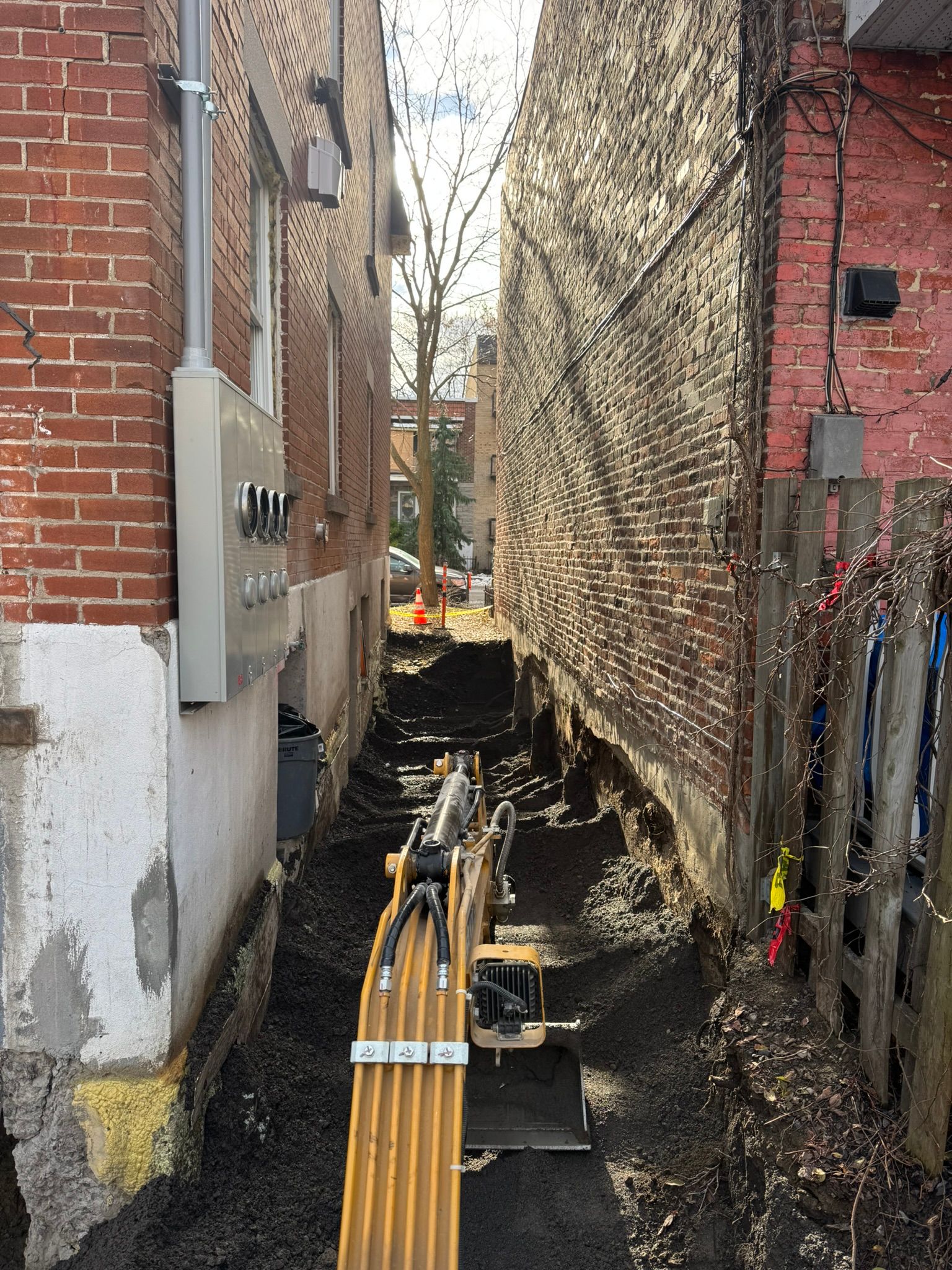 Chantier étroit entre deux bâtiments en briques. Une excavatrice est en activité dans une tranchée.
