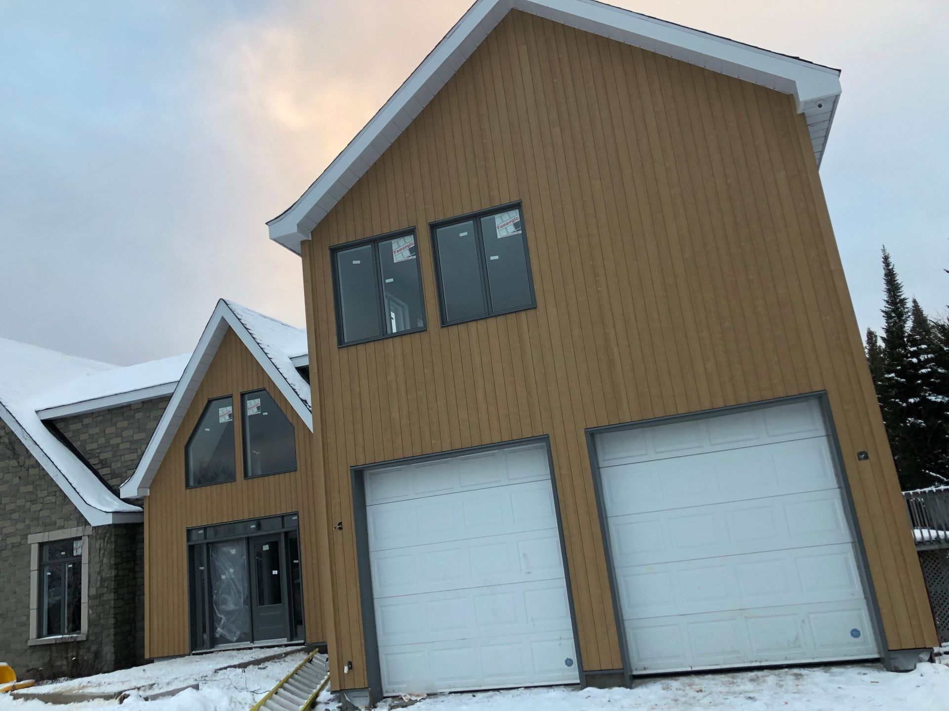 Extérieur de la maison avec bardage beige et portes de garage blanches, neige au sol, ciel nuageux.
