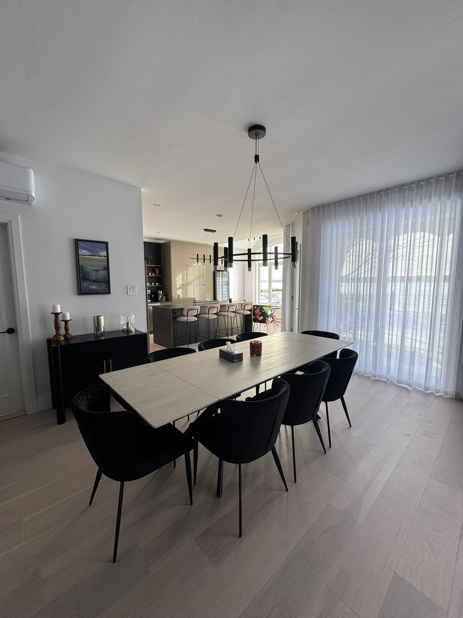 Salle à manger avec grande table, chaises noires et luminaire moderne. Parquet clair et murs blancs.
