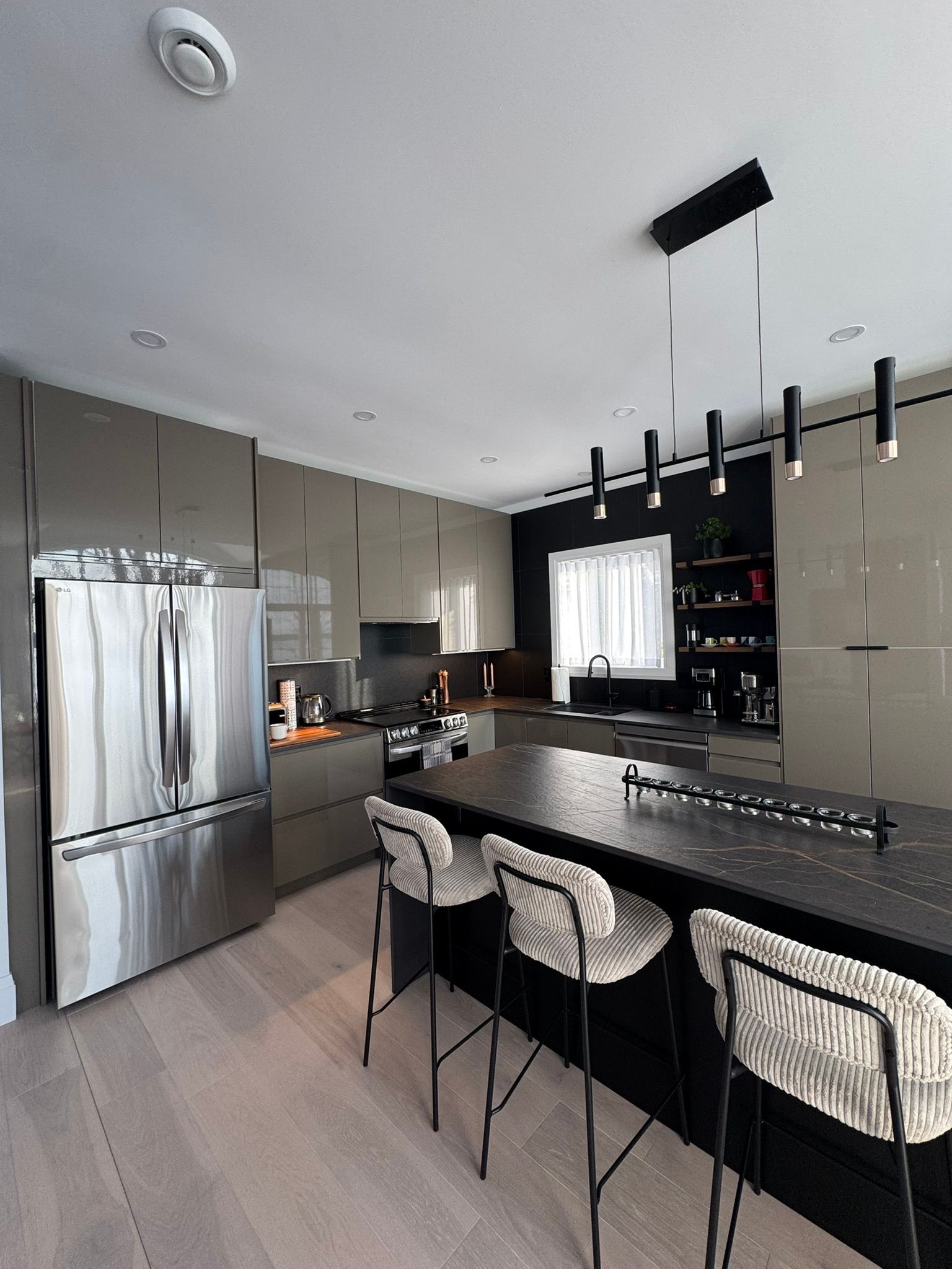 Cuisine moderne avec appareils électroménagers en acier inoxydable, îlot central foncé et trois tabourets de bar.