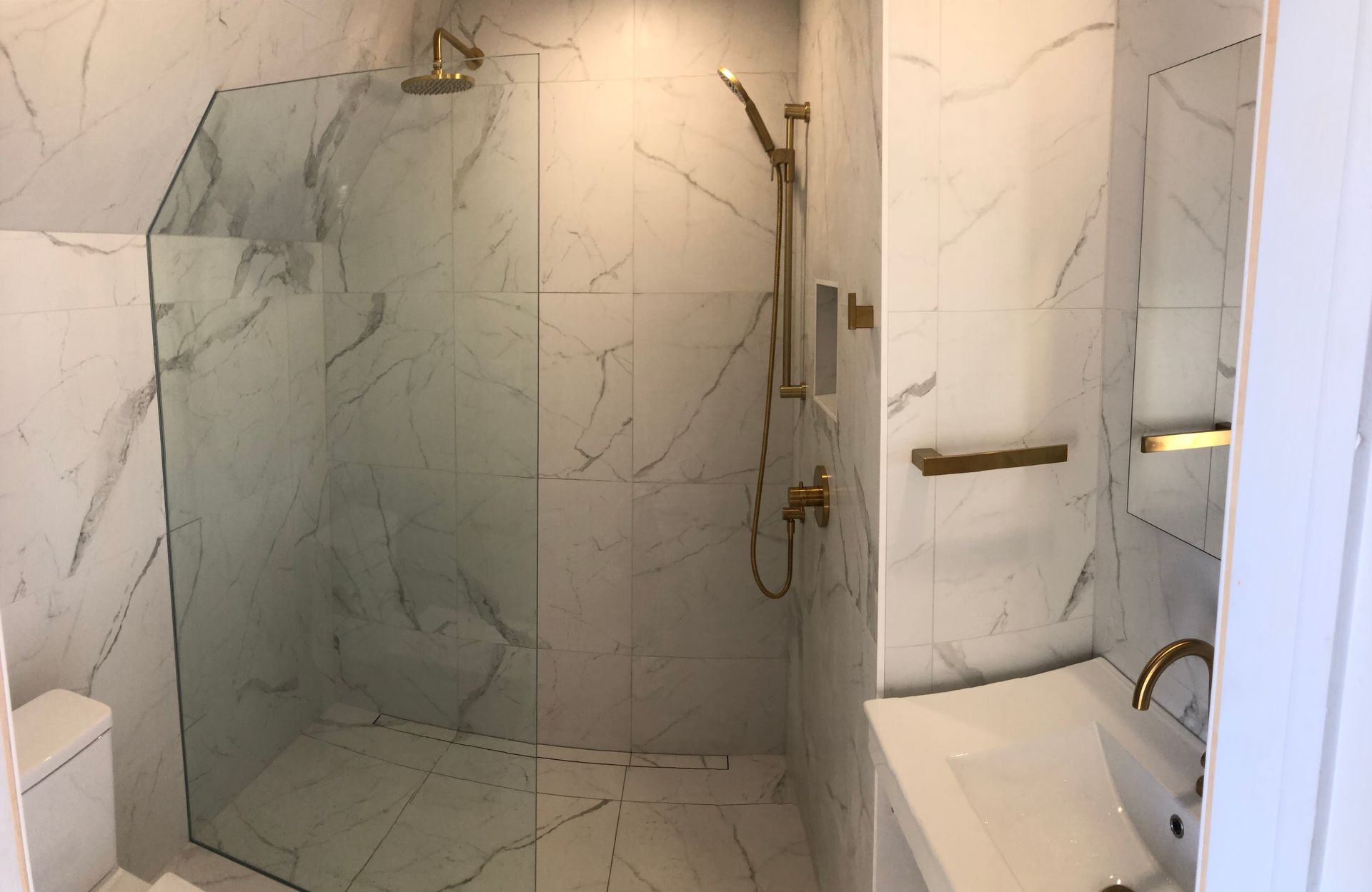 Salle de bains avec carrelage en marbre blanc, robinetterie dorée, cabine de douche en verre et lavabo blanc.