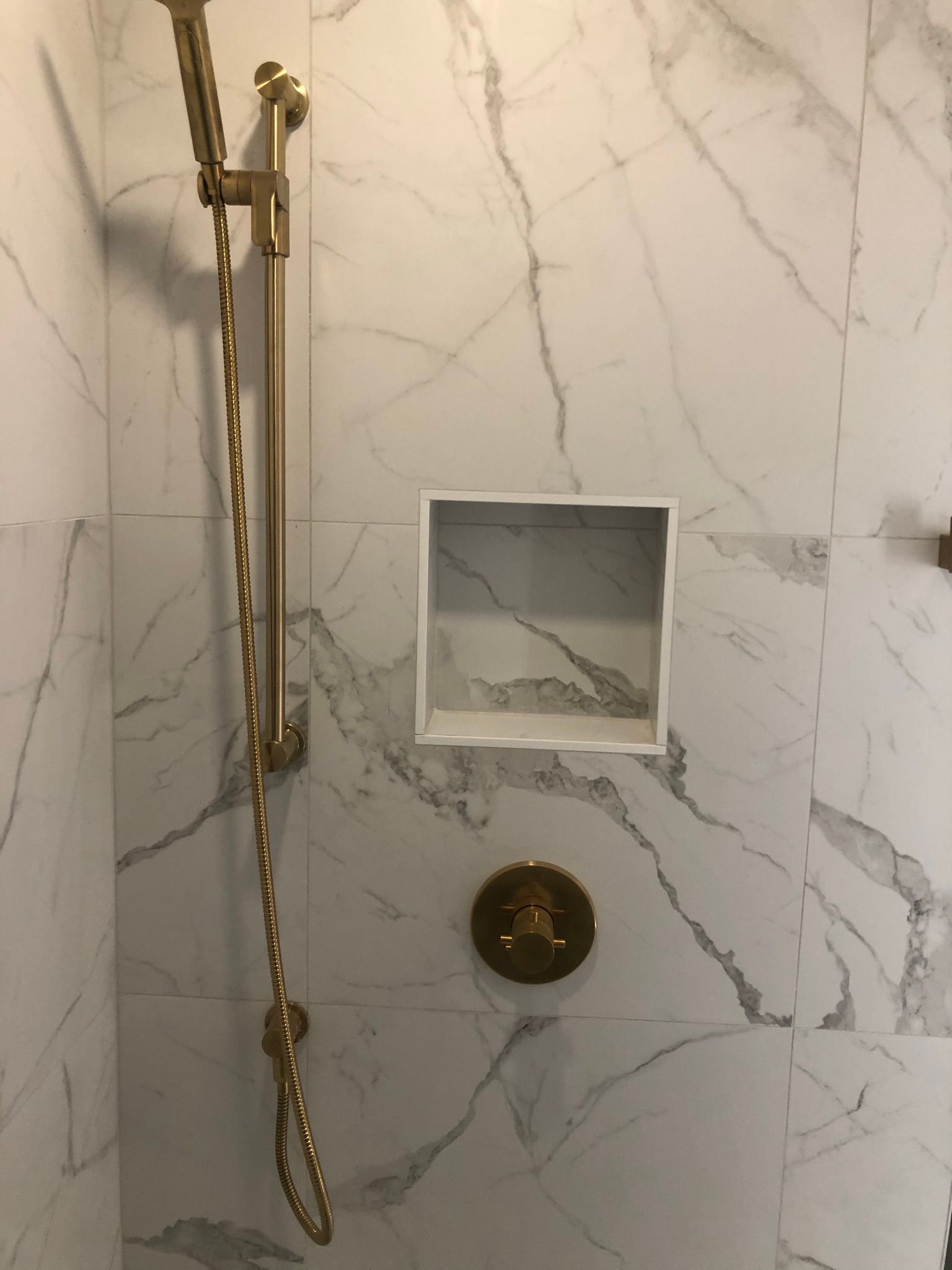 Robinetterie de douche dorée sur un mur carrelé de marbre blanc avec une étagère intégrée.