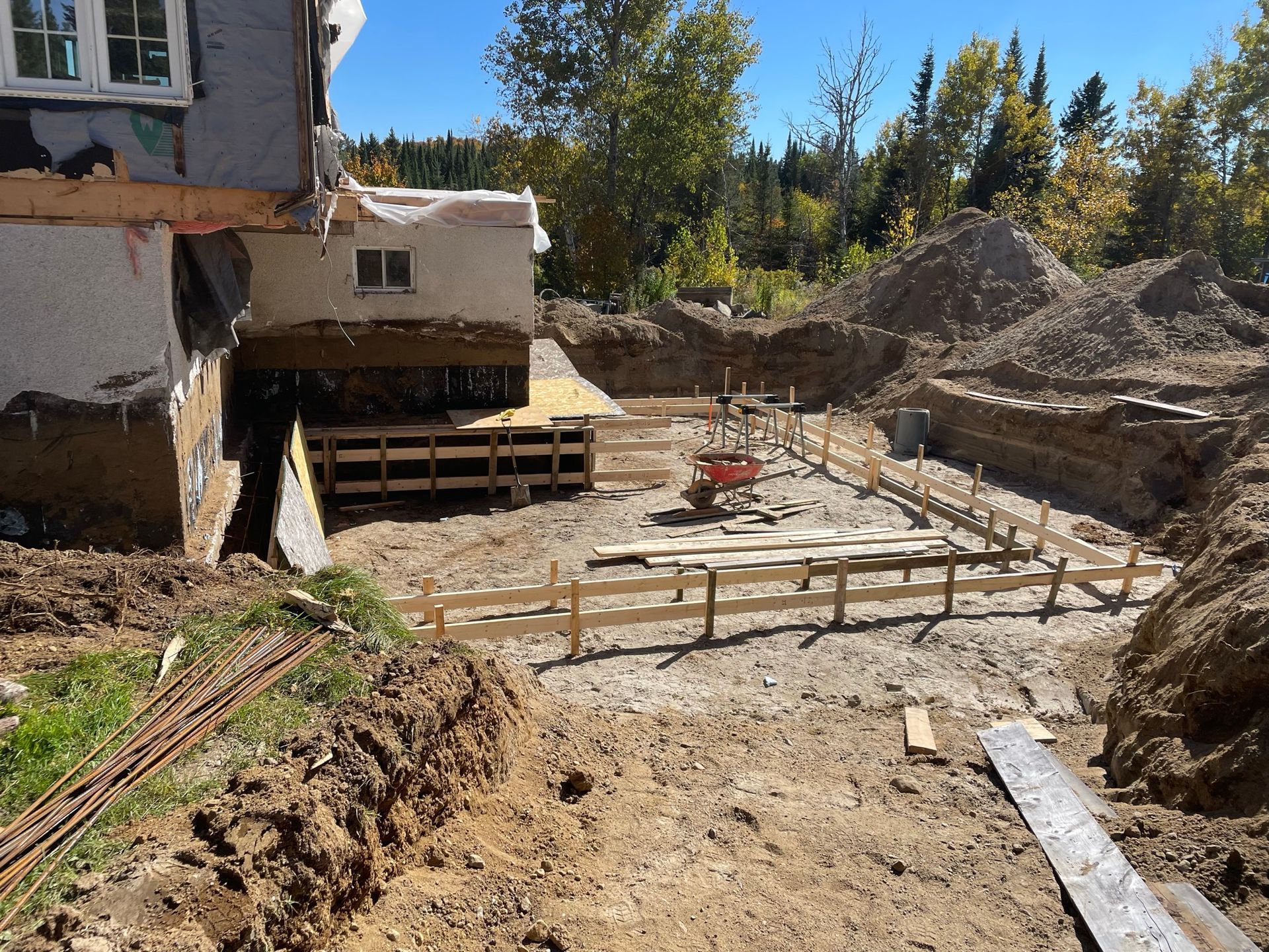 Chantier : coffrages de fondation, terre excavée et bâtiment existant.
