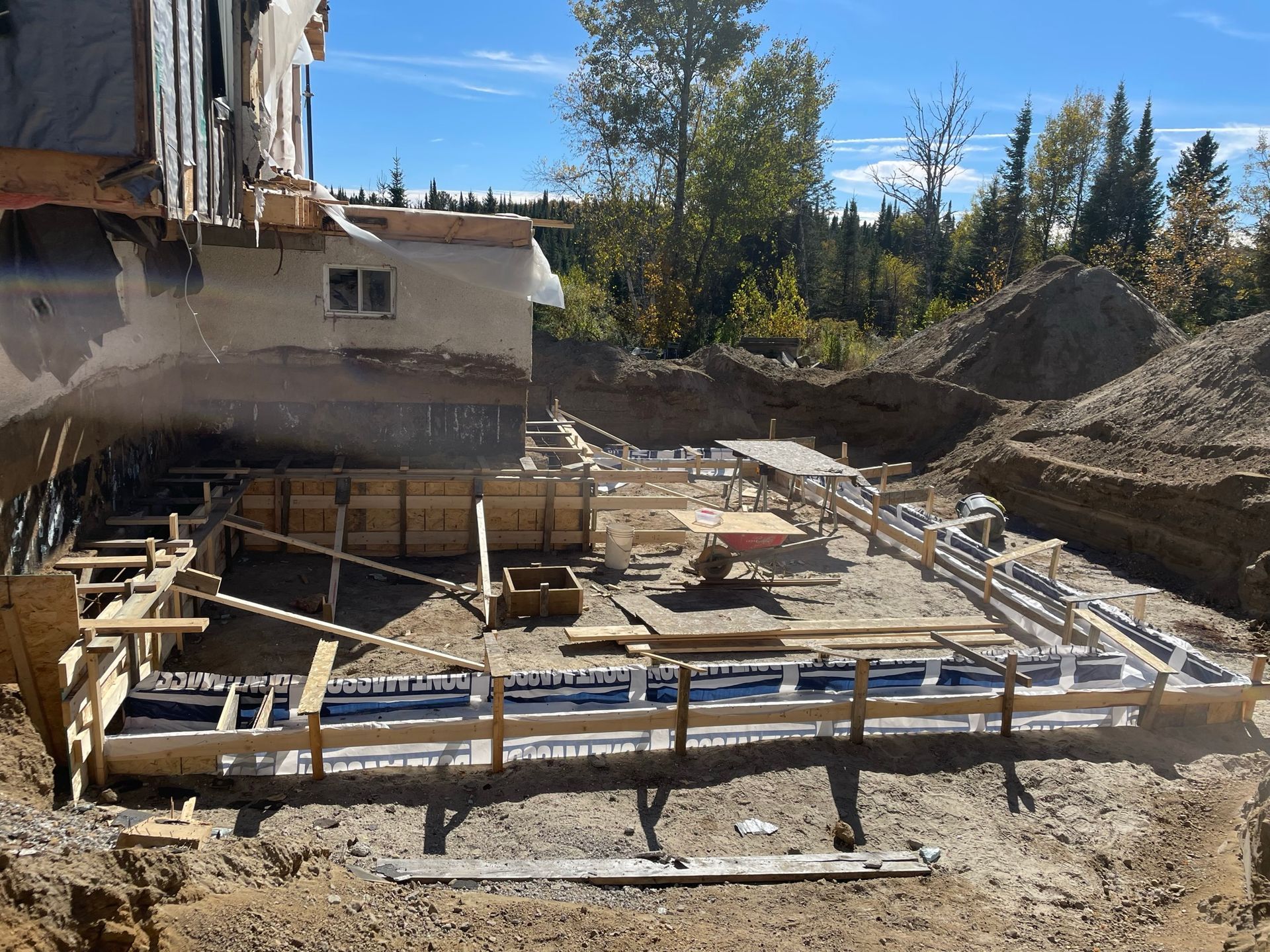 Chantier de construction avec coffrages en bois pour une fondation en béton à côté d'un bâtiment existant.
