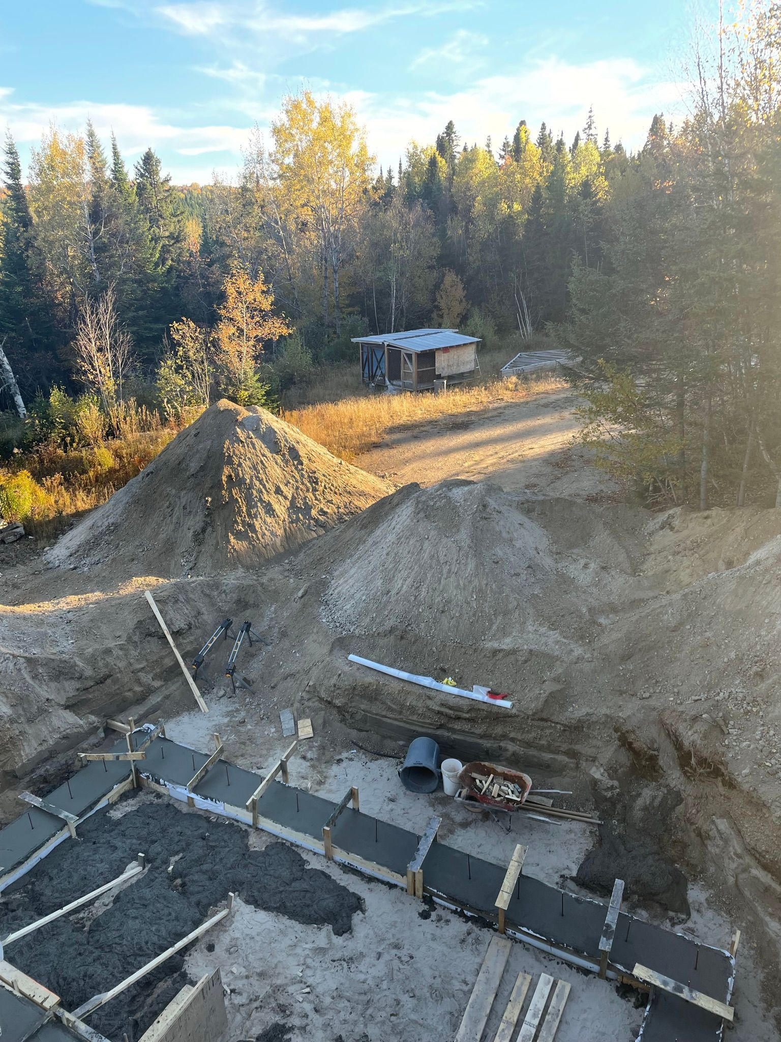 Chantier de construction avec fondations en béton et tas de terre, une petite cabane en arrière-plan.