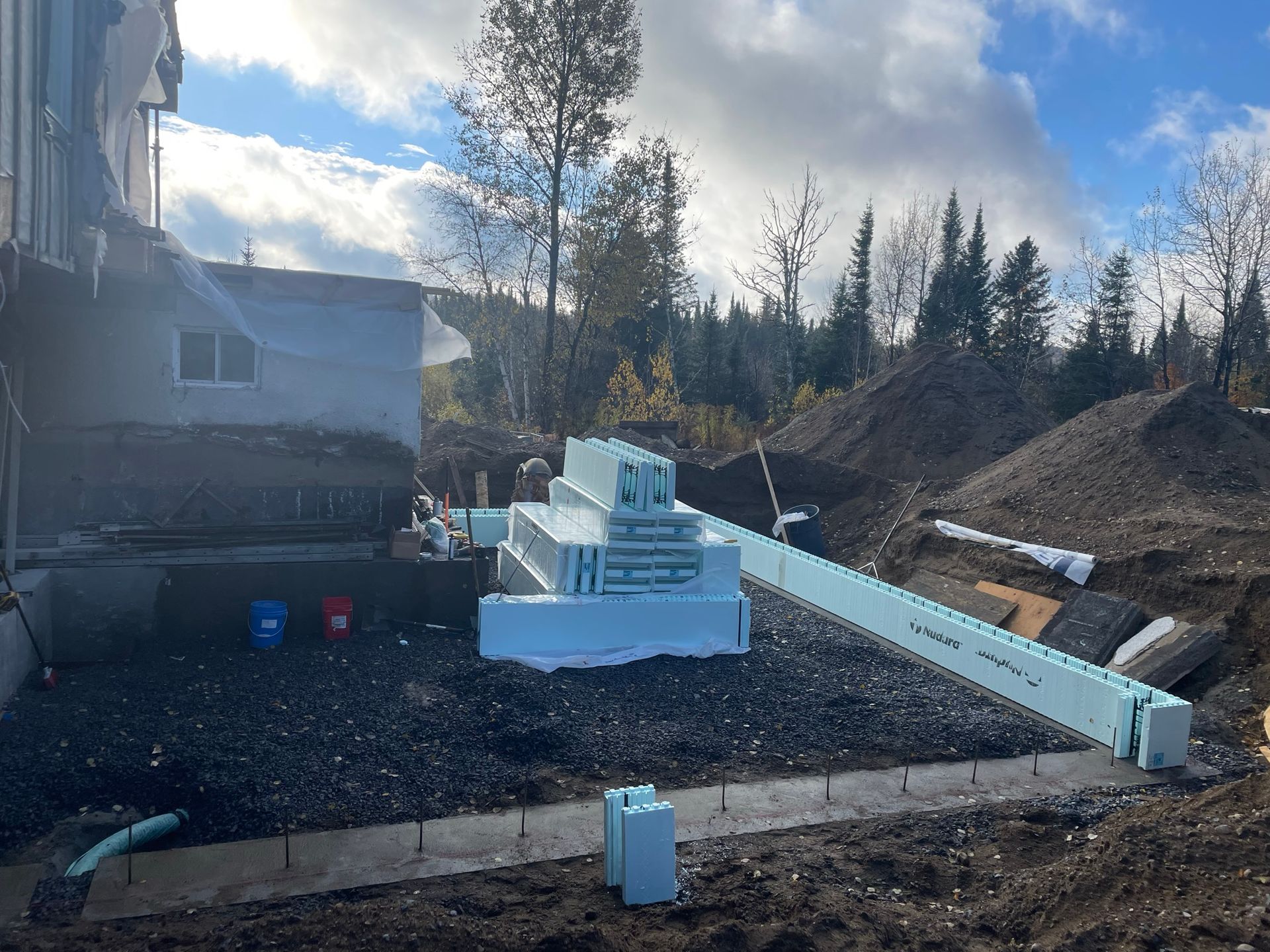 Chantier de construction avec des blocs de mousse empilés, des fondations en béton et un tas de terre sous un ciel nuageux.