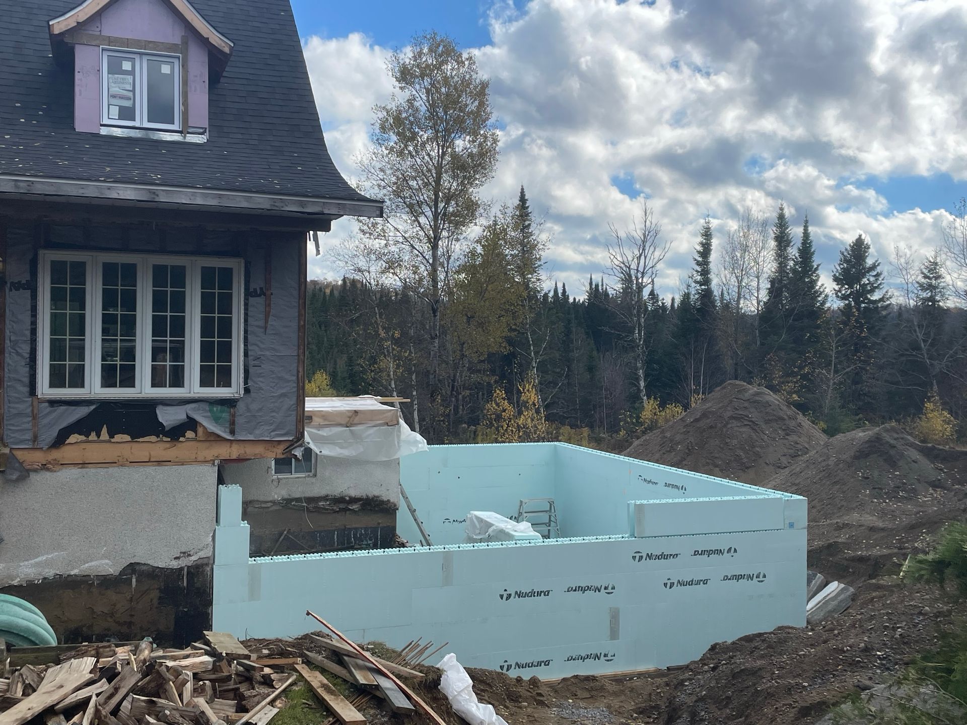 Une maison en construction, aux fondations en mousse bleu clair. Un décor boisé sous un ciel nuageux.