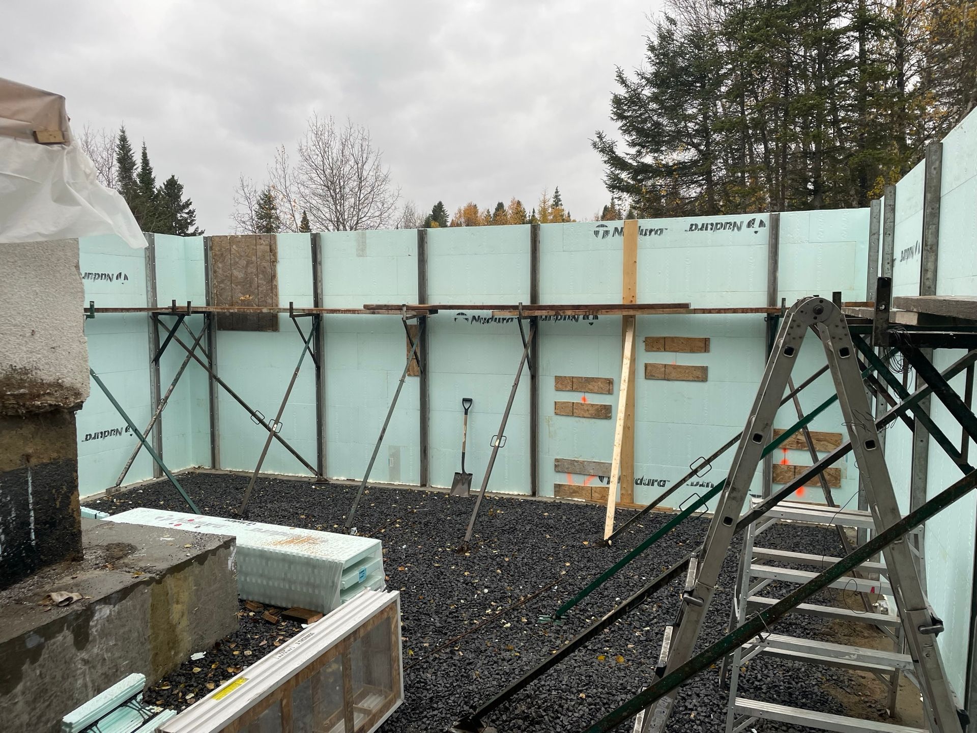 Chantier de construction avec coffrages en béton isolés, sol en gravier, supports et une échelle.
