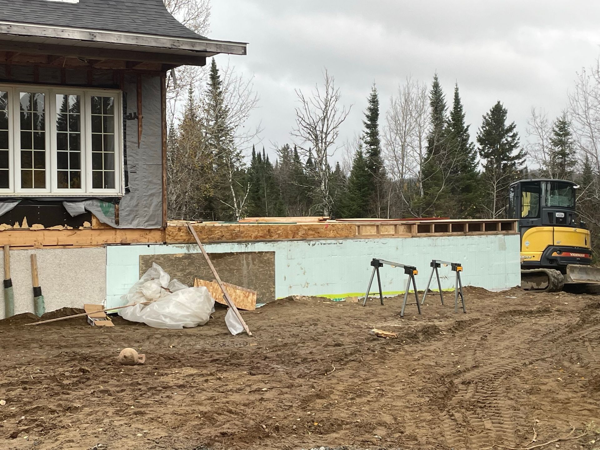 Chantier de construction avec charpente de maison, isolation et engins de terrassement. Temps couvert.
