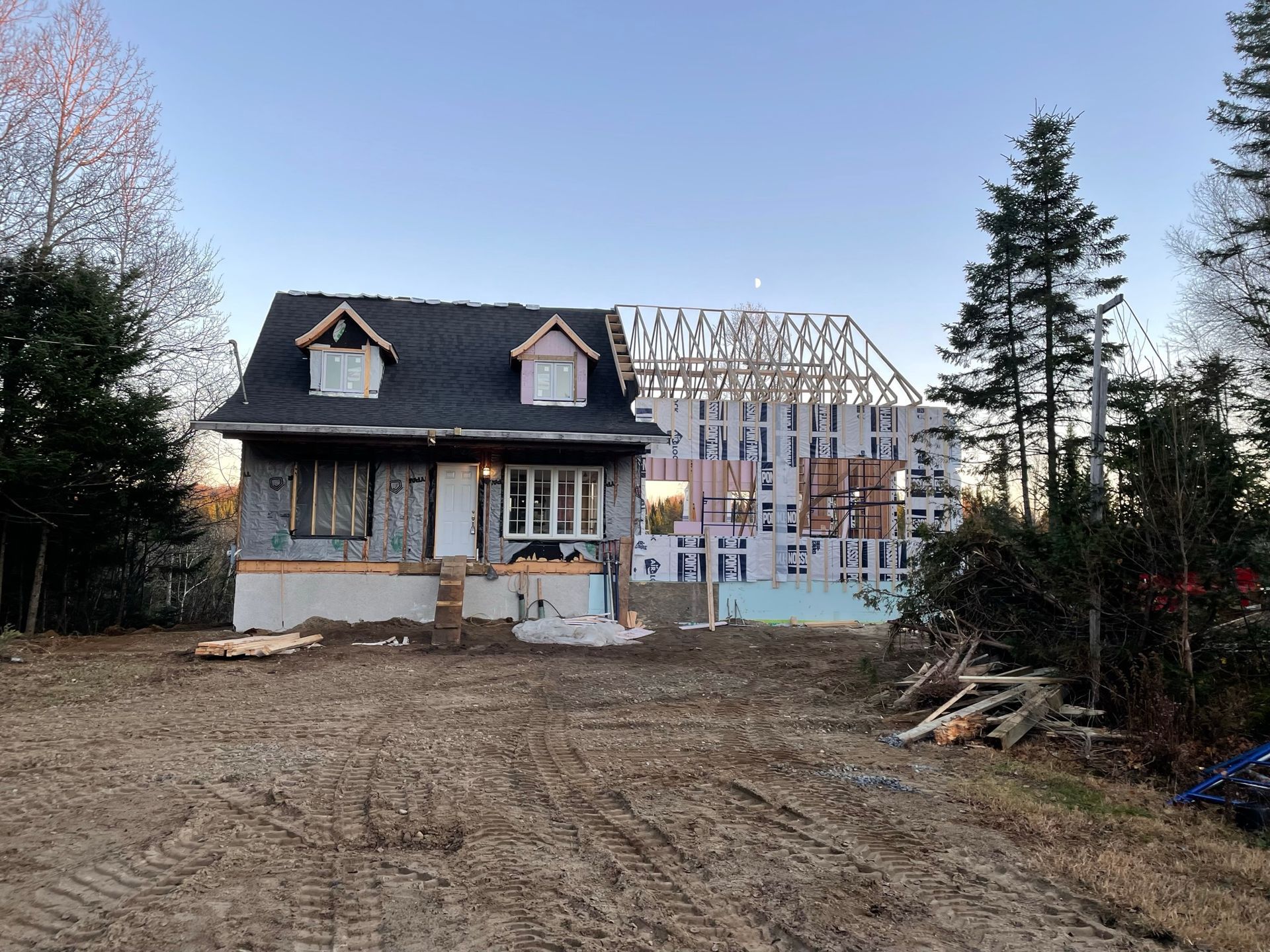 Maison en construction ; structure existante au premier plan, nouvelle charpente à l'arrière-plan ; entourée d'arbres et de terre.