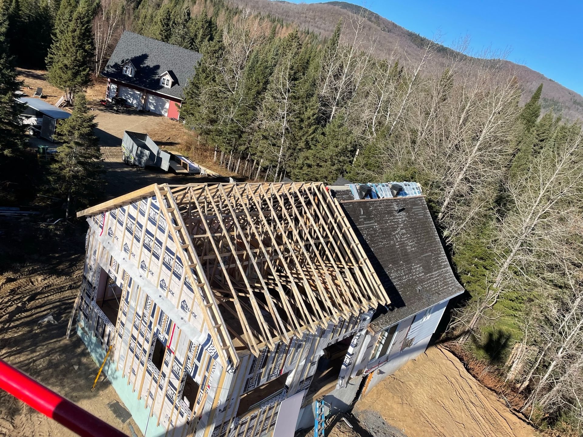 Chantier : maison en construction à ossature bois apparente, nichée à flanc de colline boisée, par une journée ensoleillée.