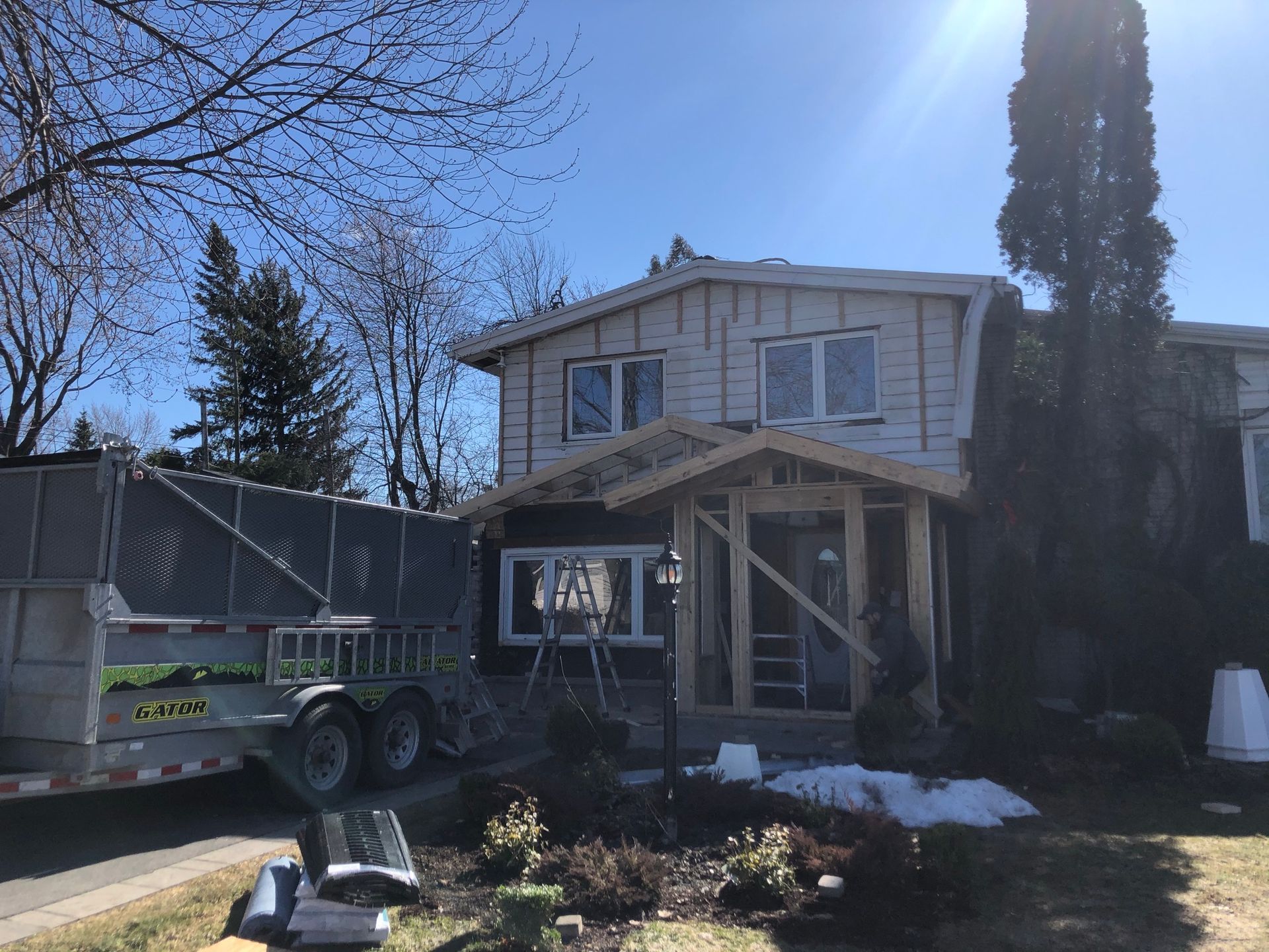 Maison en construction avec charpente apparente, journée ensoleillée. Présence d'une caravane et de neige.