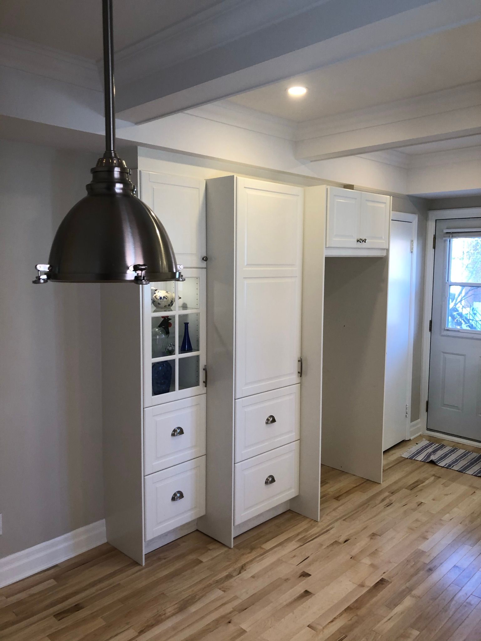 Cuisine avec placards blancs, sol en bois, une grande suspension lumineuse argentée et une porte sur la droite.