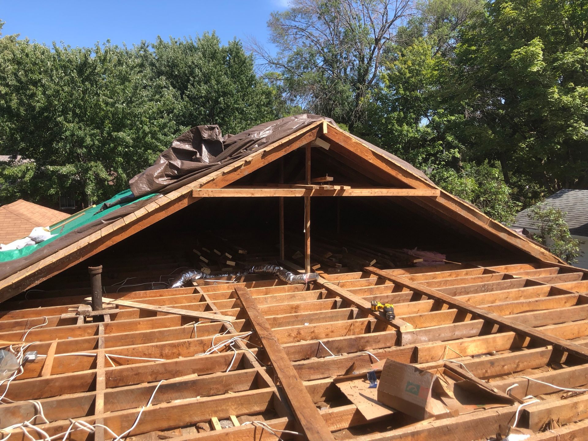 Charpente du toit apparente lors des travaux de rénovation, avec poutres en bois, ciel bleu et arbres en arrière-plan.