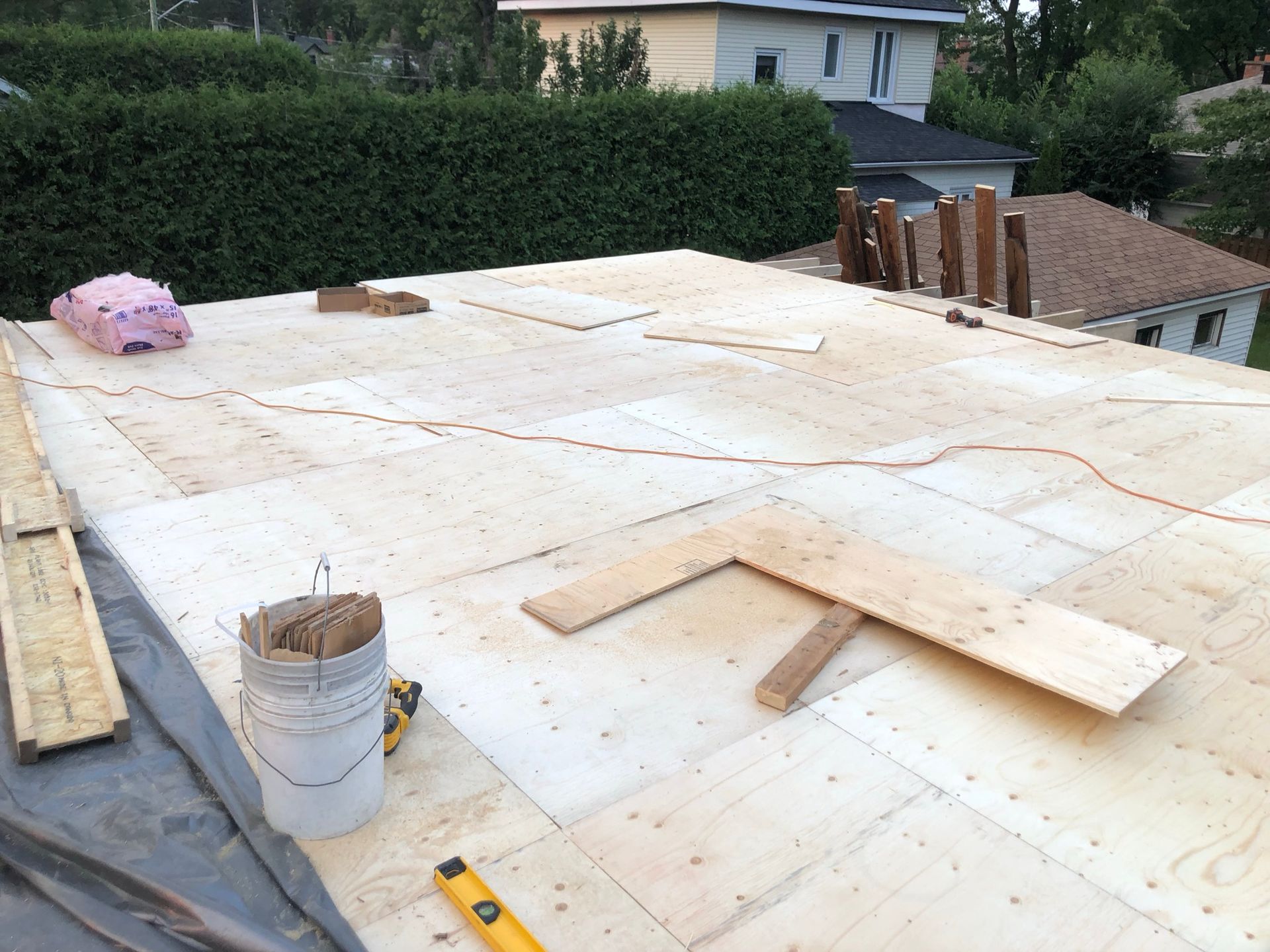 Toit recouvert de contreplaqué, outils de construction visibles et une maison en arrière-plan.