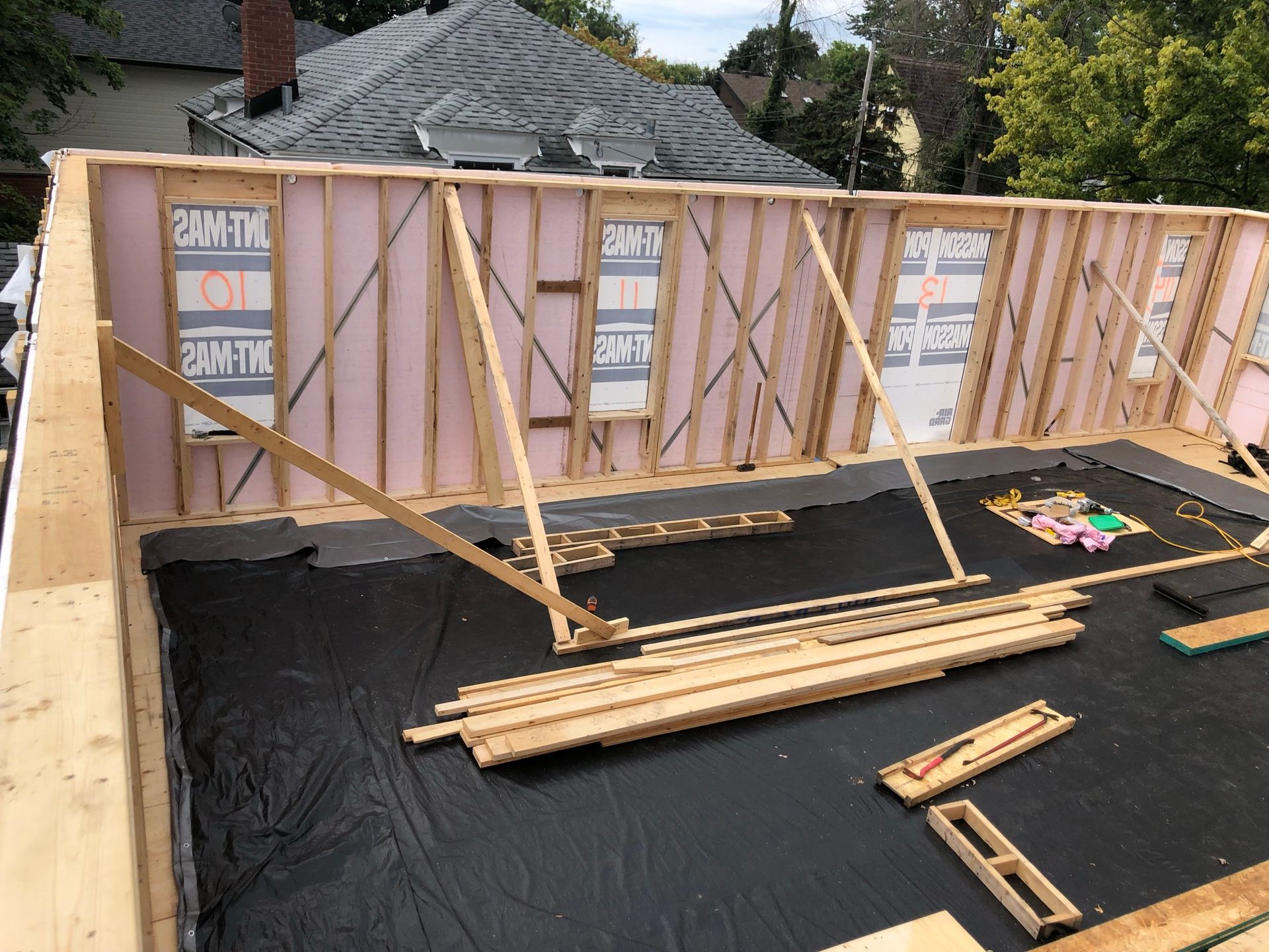 Chantier : Ossature en bois avec isolation rose, supports temporaires et sol recouvert d'une bâche noire.