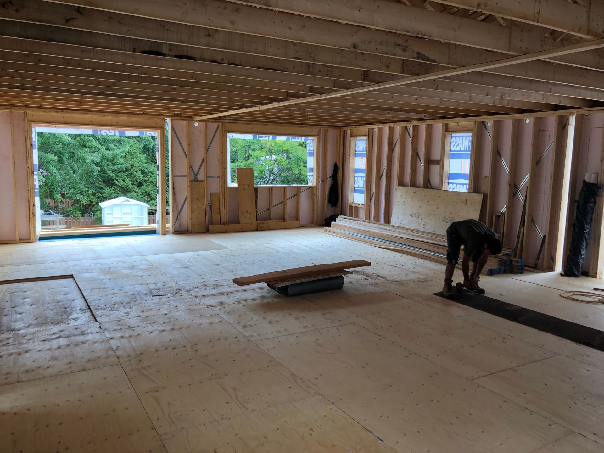 Intérieur d'une pièce en construction avec une charpente en bois apparente et un ouvrier installant un revêtement de sol.