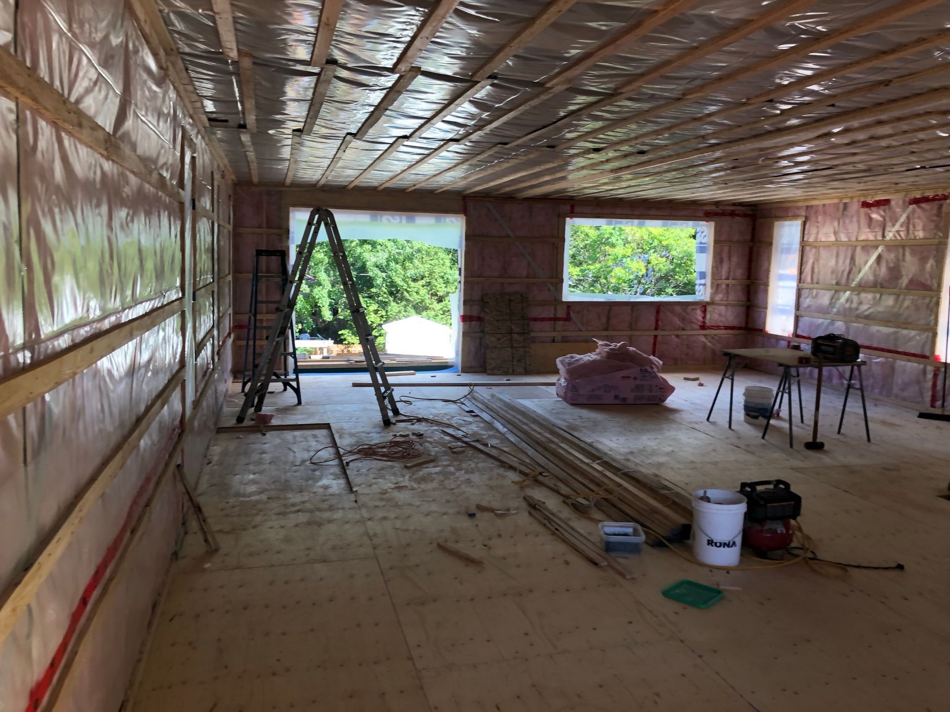 Intérieur d'un bâtiment en construction, montrant l'isolation des murs et du plafond, une échelle et une vue extérieure.