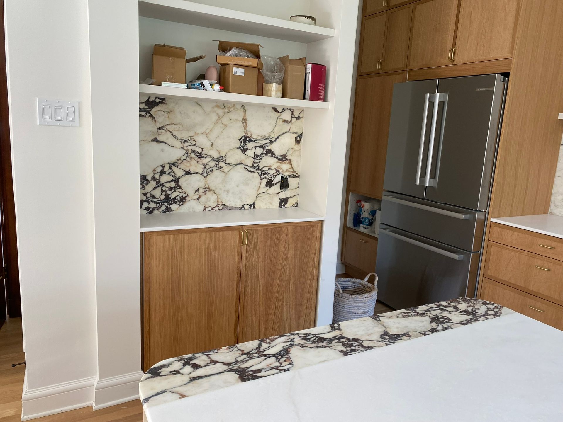 Cuisine avec placards en bois clair, étagères intégrées, crédence en marbre, réfrigérateur en acier inoxydable et plan de travail blanc.